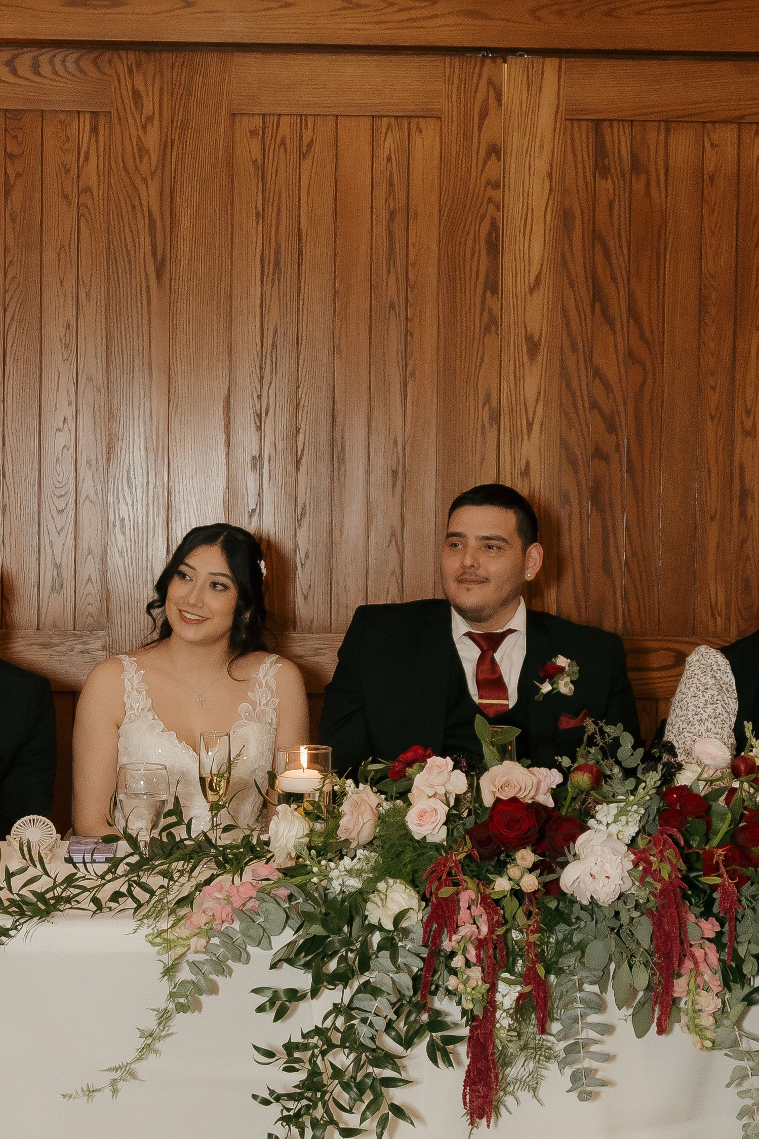 Smiling bride and groom sit at sweetheart table adorned with lush floral arrangements and candles.