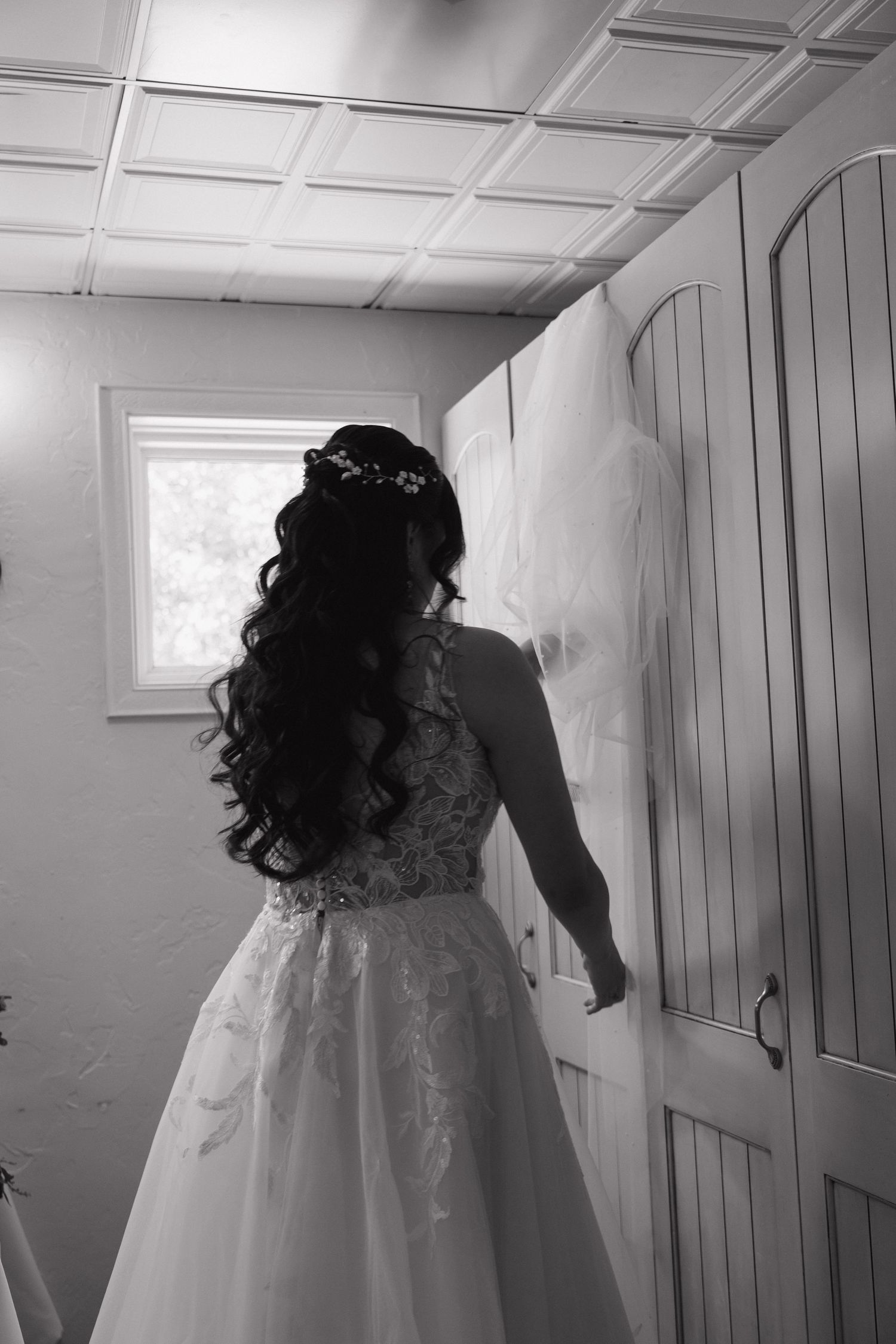 Bride in lace wedding gown with long veil and floral hair accessory standing by a door.