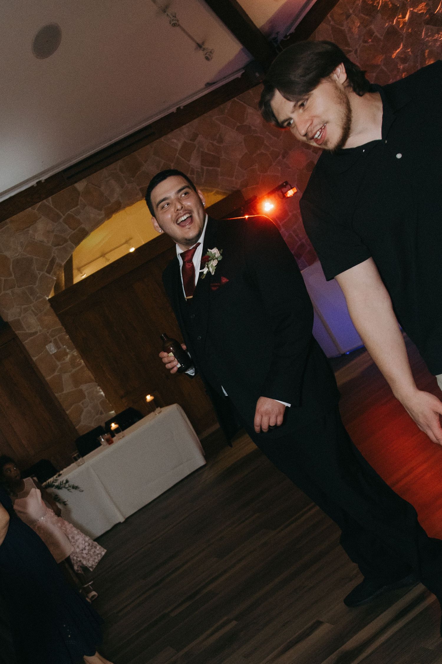 Groom in black suit laughing and dancing with guest at wedding reception.