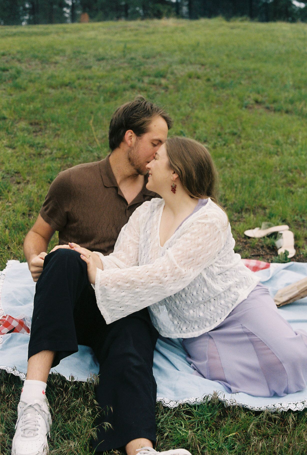 A couple shares a tender kiss while sitting on a picnic blanket in a green field.