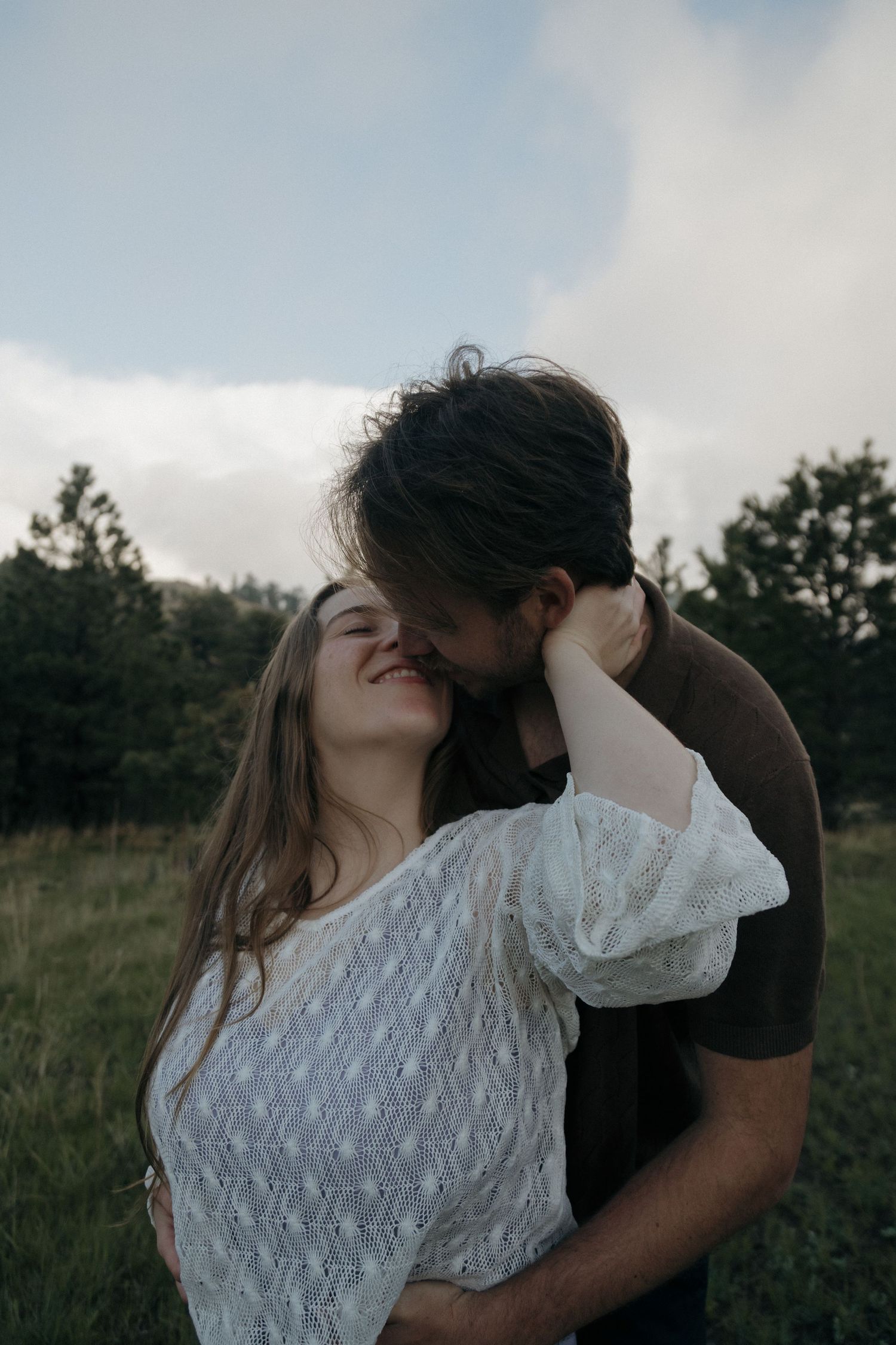 Romantic couple embracing and kissing in a green meadow with pine trees and cloudy sky backdrop.