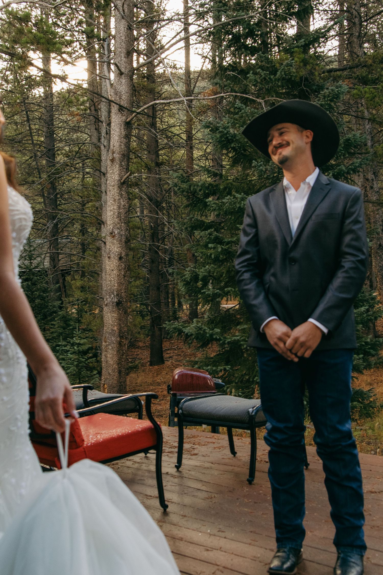 A groom in a black suit and cowboy hat smiles during an outdoor forest wedding ceremony.