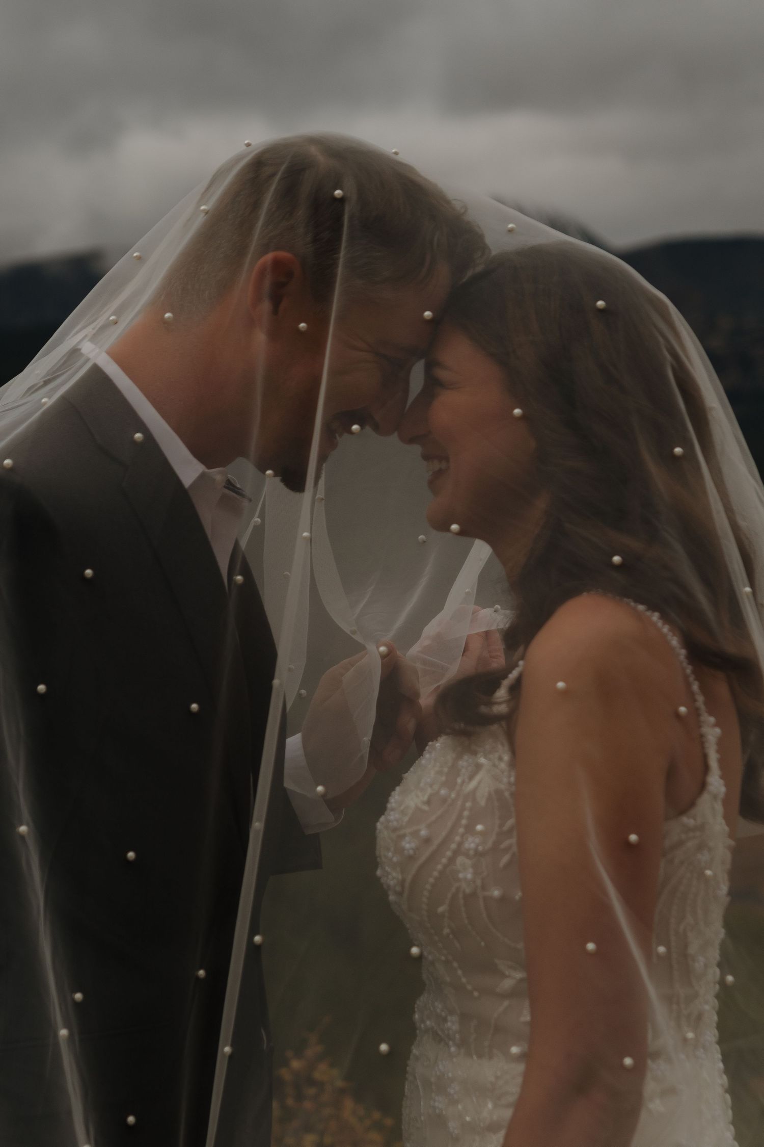A romantic wedding photo of a couple sharing an intimate moment under a veil against a moody sky.