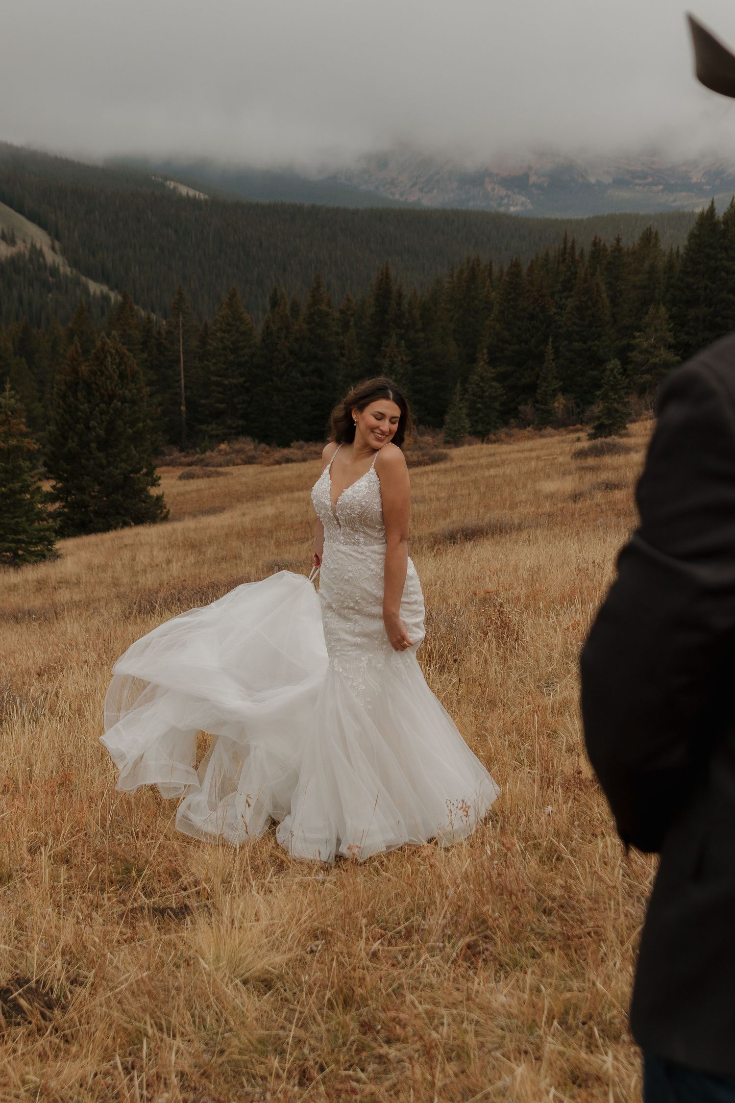 A bride in a flowing white dress twirls in a mountain meadow with golden grass and evergreen trees.