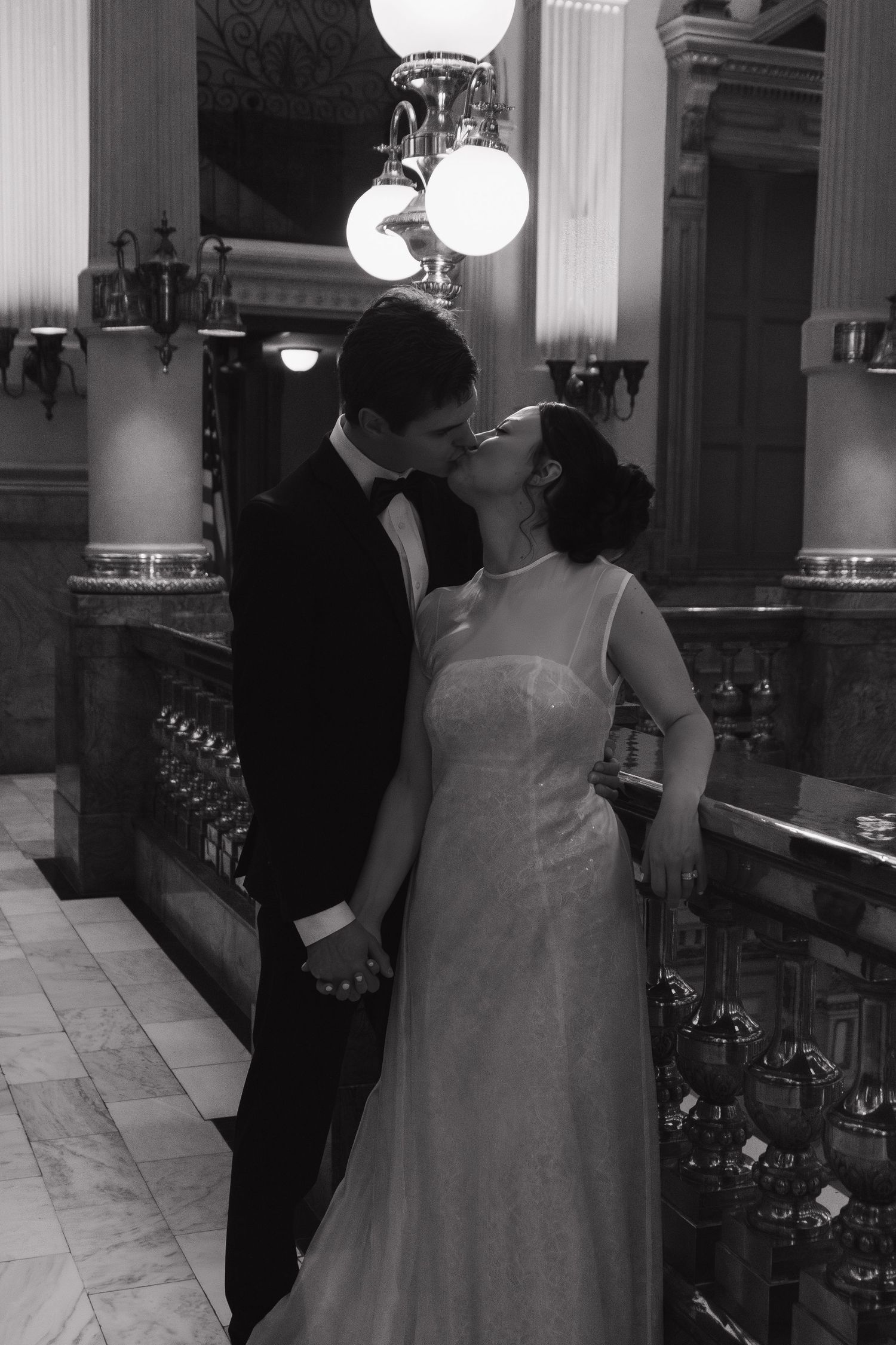 A black and white wedding photograph capturing a romantic kiss beneath vintage light fixtures.
