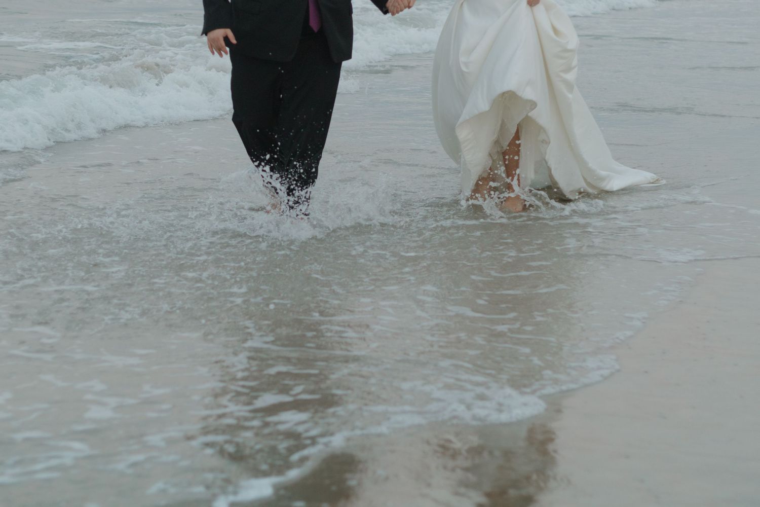 A couple walks hand in hand through shallow ocean water, with the bride's white wedding dress trailing behind them.