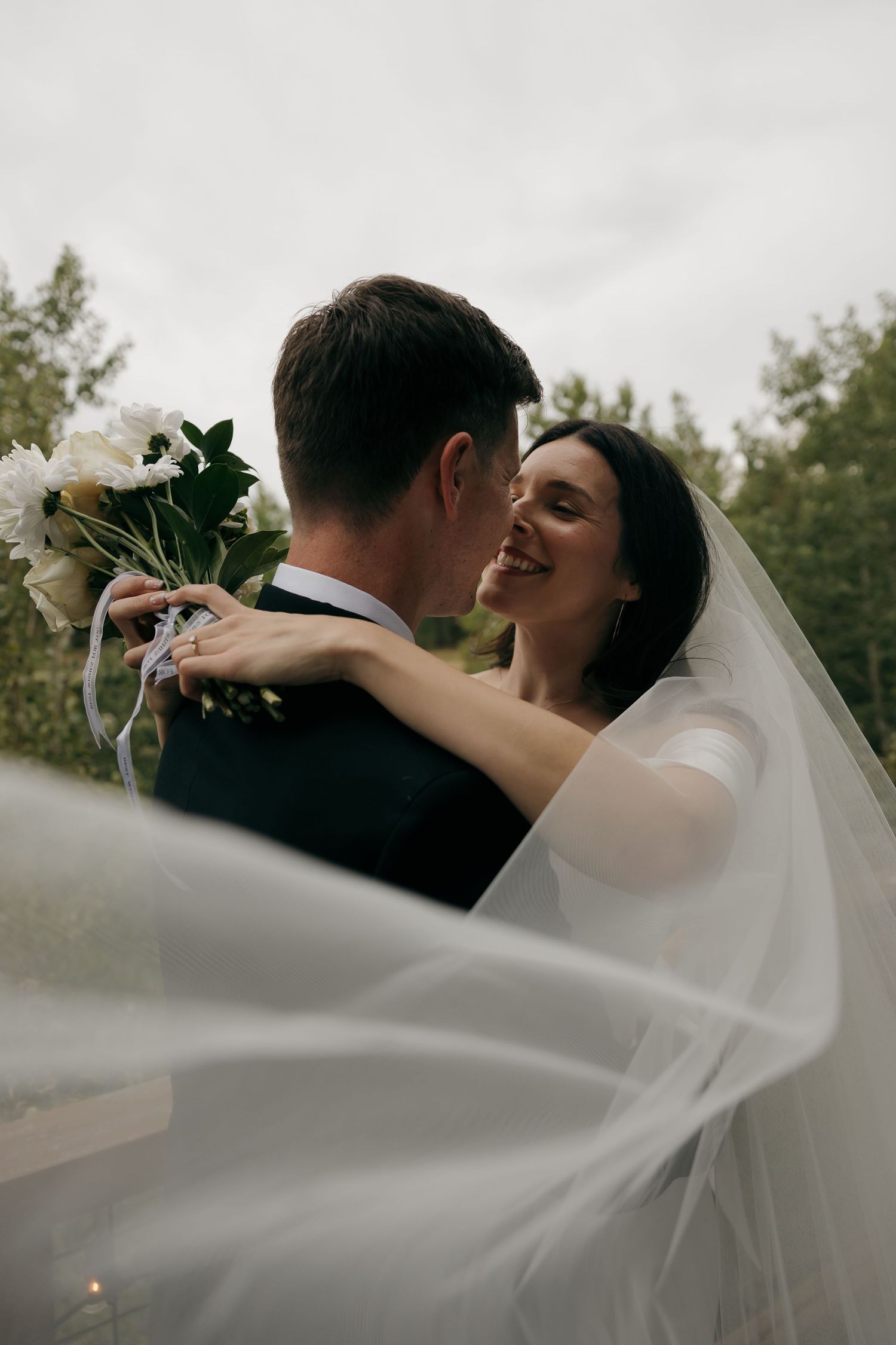 Smiling bride embracing groom with white peony bouquet, veil flowing in the wind outdoors.