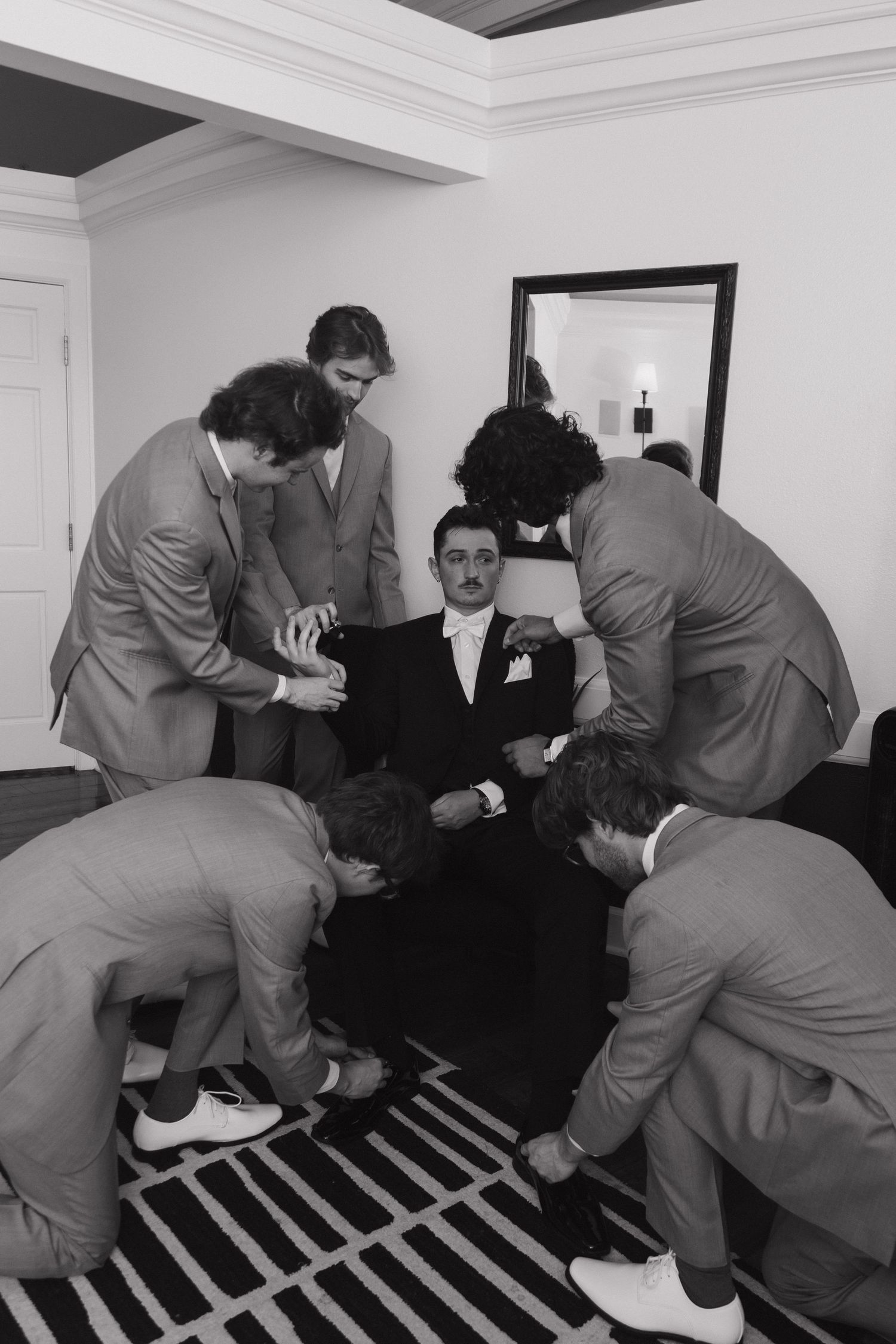 Groomsmen in suits helping groom get ready, surrounding him in chair, black and white wedding photo.