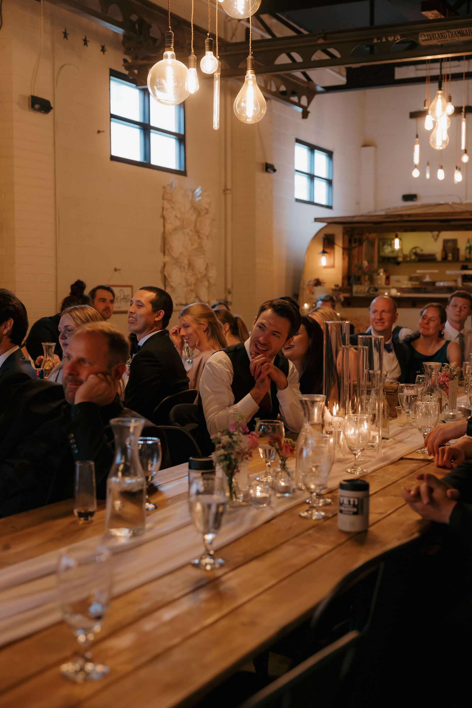 Wedding guests laughing and enjoying dinner at long wooden table with candles and Edison bulb lighting.