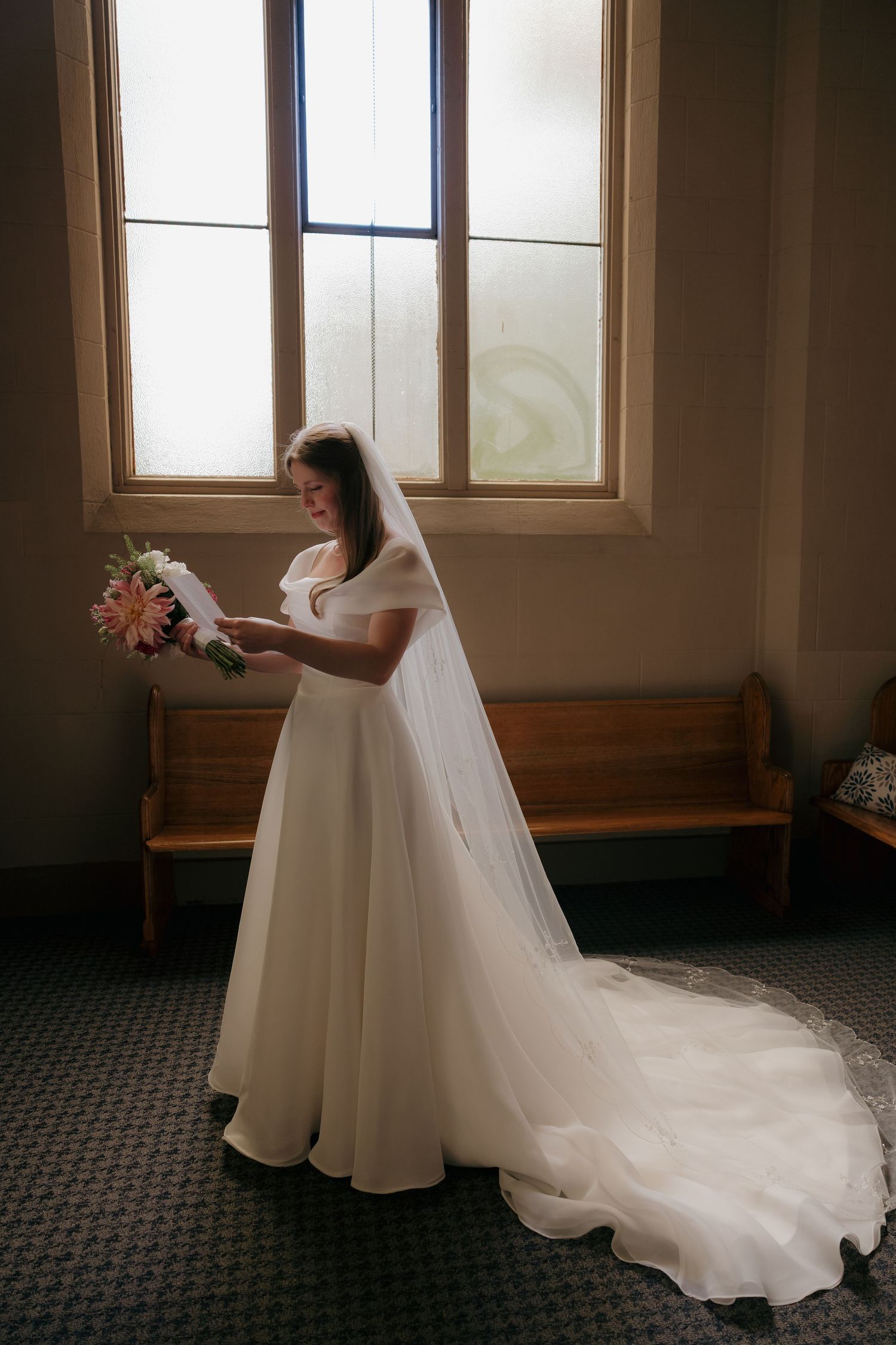 Bride in white wedding gown with veil holds bouquet by church window.