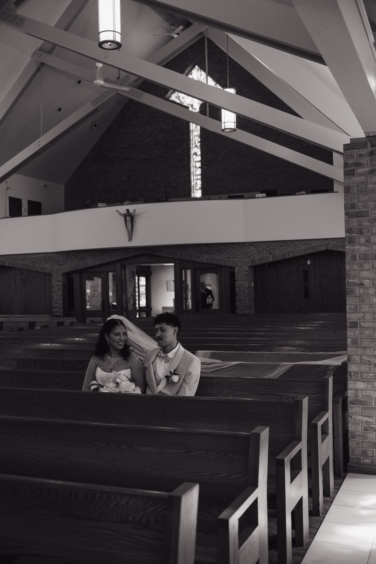 Black and white photo of a bride and groom sharing an intimate moment seated in church pews during their wedding ceremony.