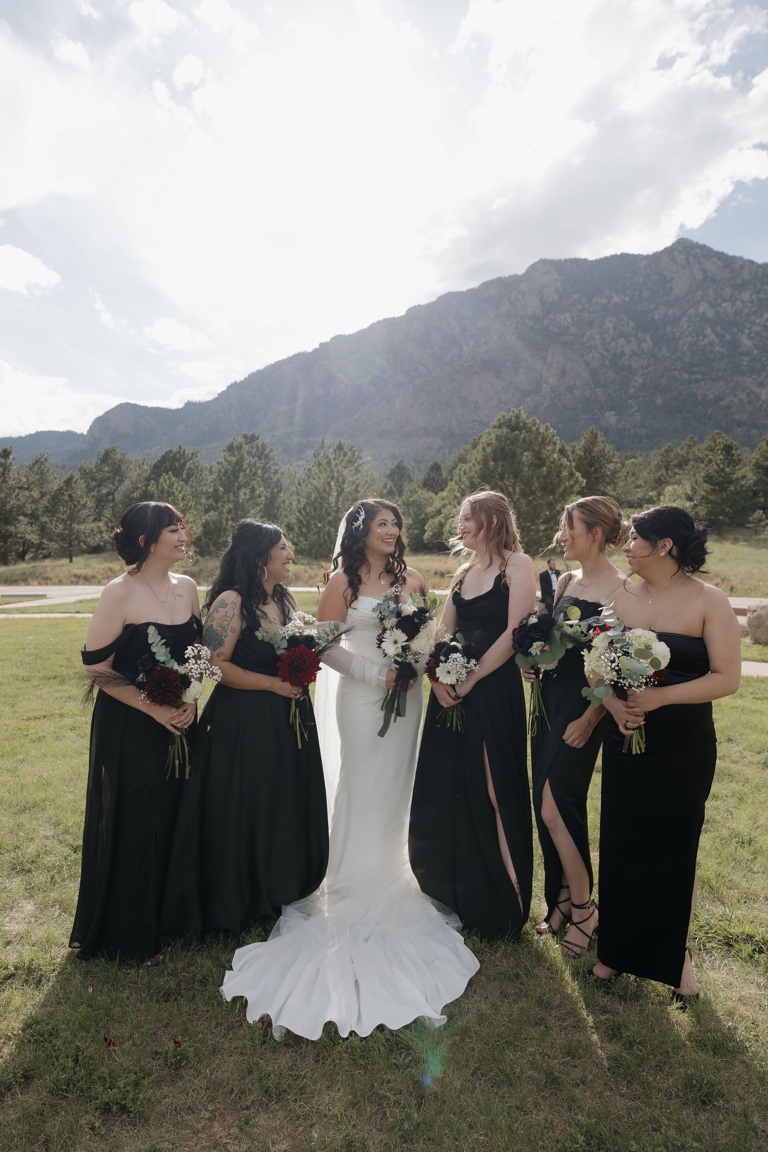 Bride in white gown with four bridesmaids in black dresses holding bouquets, posing in front of mountain scenery.