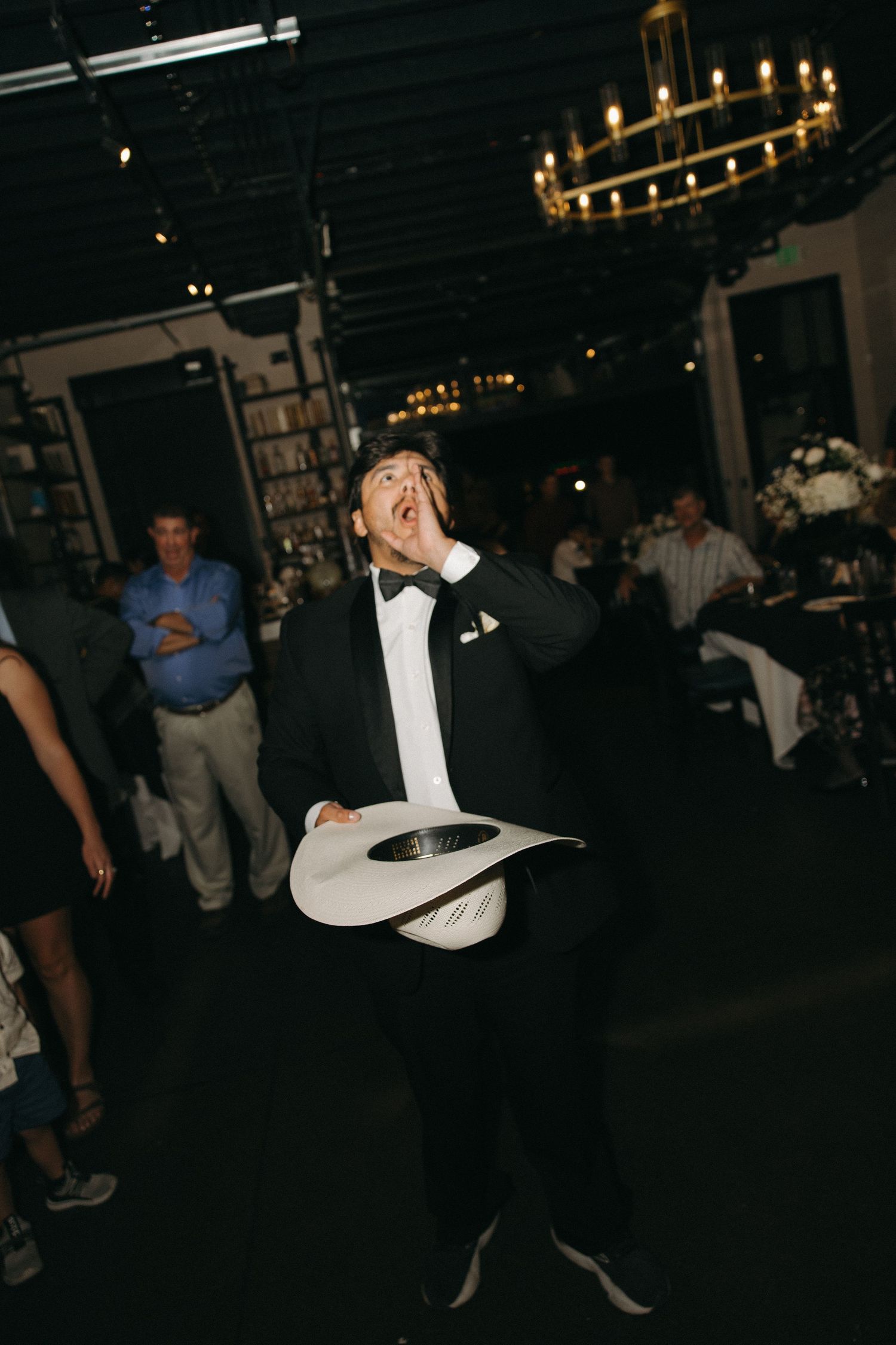 Man in tuxedo energetically holds white cowboy hat at dark indoor wedding reception venue.