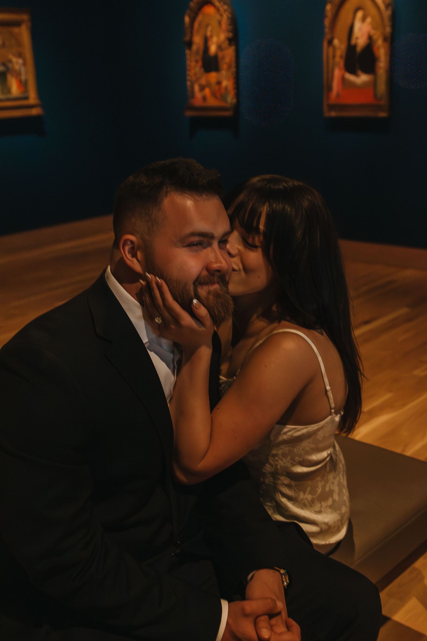 A romantic couple shares intimate kisses in a dimly lit art gallery with paintings visible on the wall.