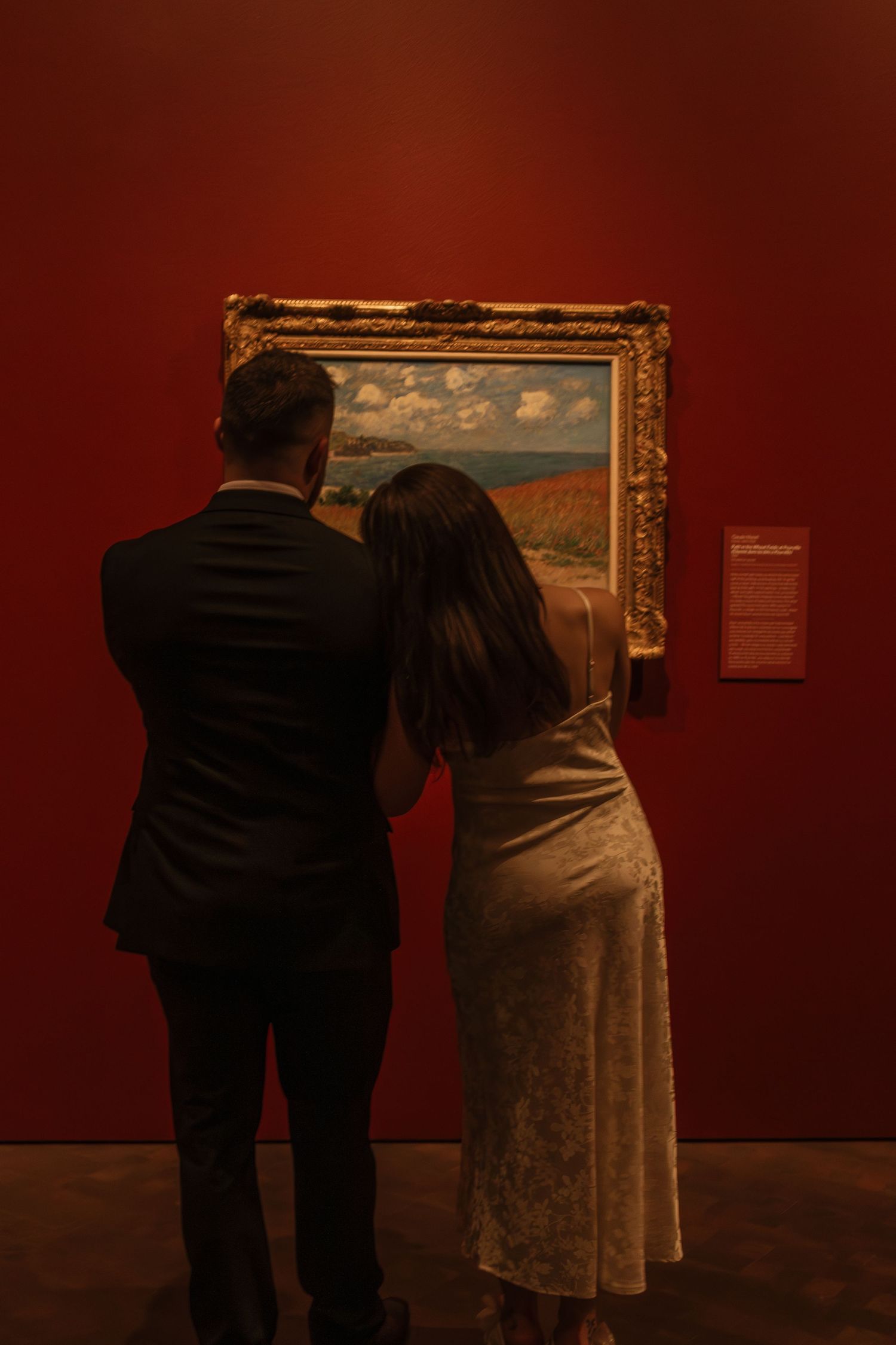 A couple admires a framed landscape painting in a museum with deep red walls.
