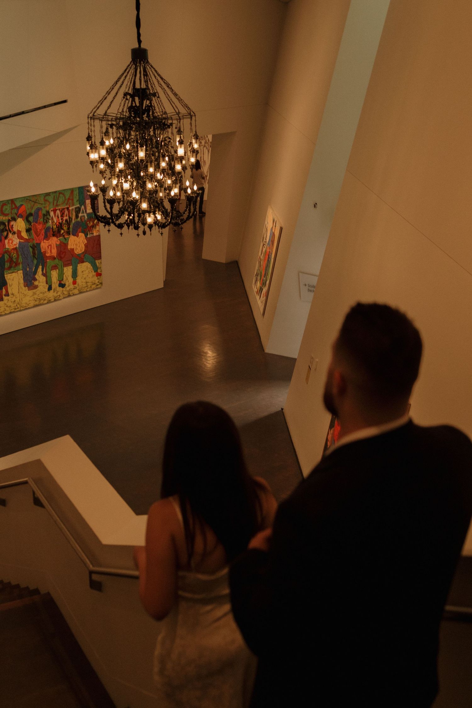 A couple embraces in a dimly lit gallery space with decorative lighting fixtures overhead.