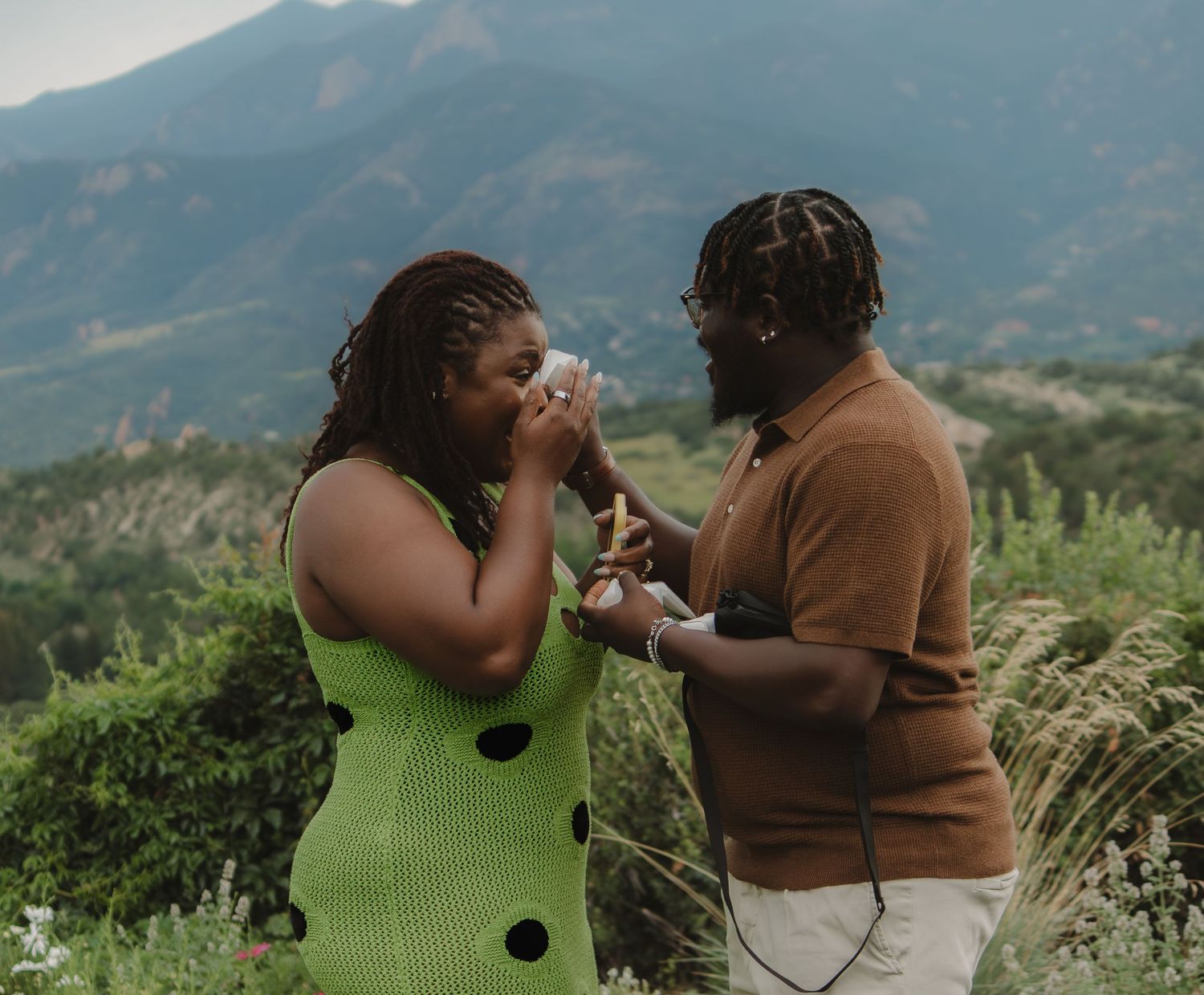Couple shares an emotional moment against a scenic mountain backdrop while wearing green and brown sweaters.