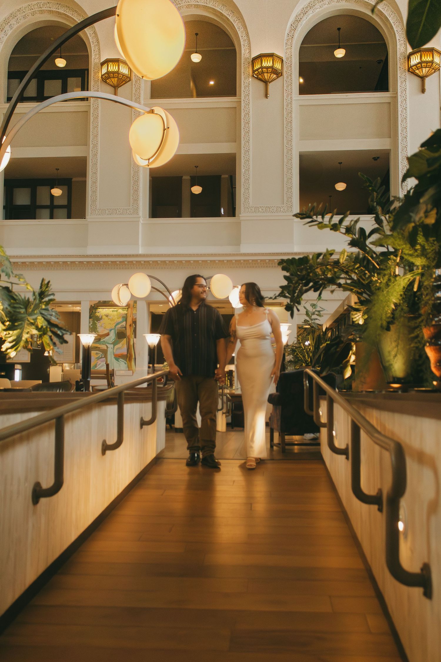 A couple walks along an elegant indoor walkway with hanging lanterns and arched architecture.