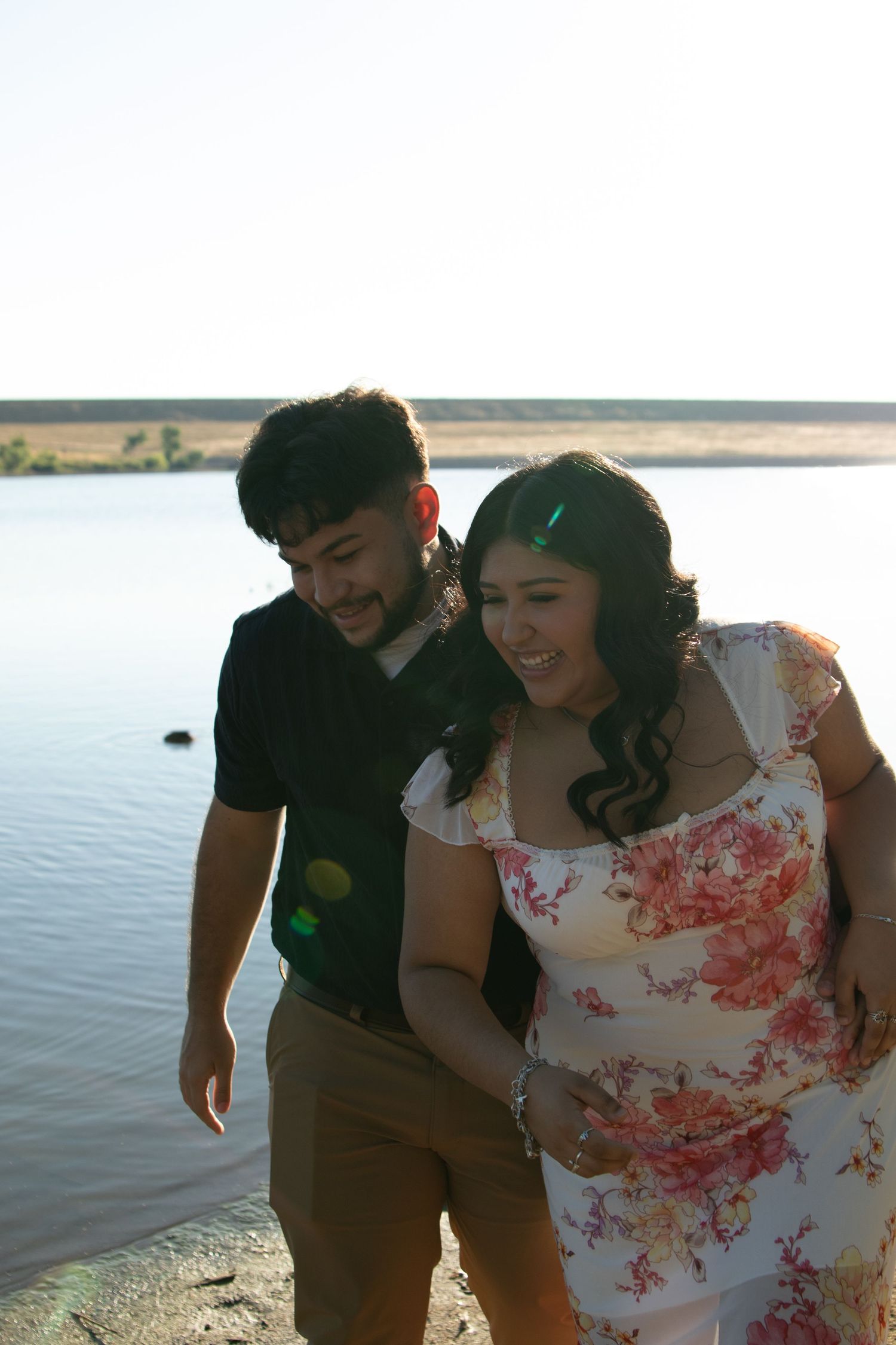 A couple shares a joyful moment together by a waterfront during golden hour.