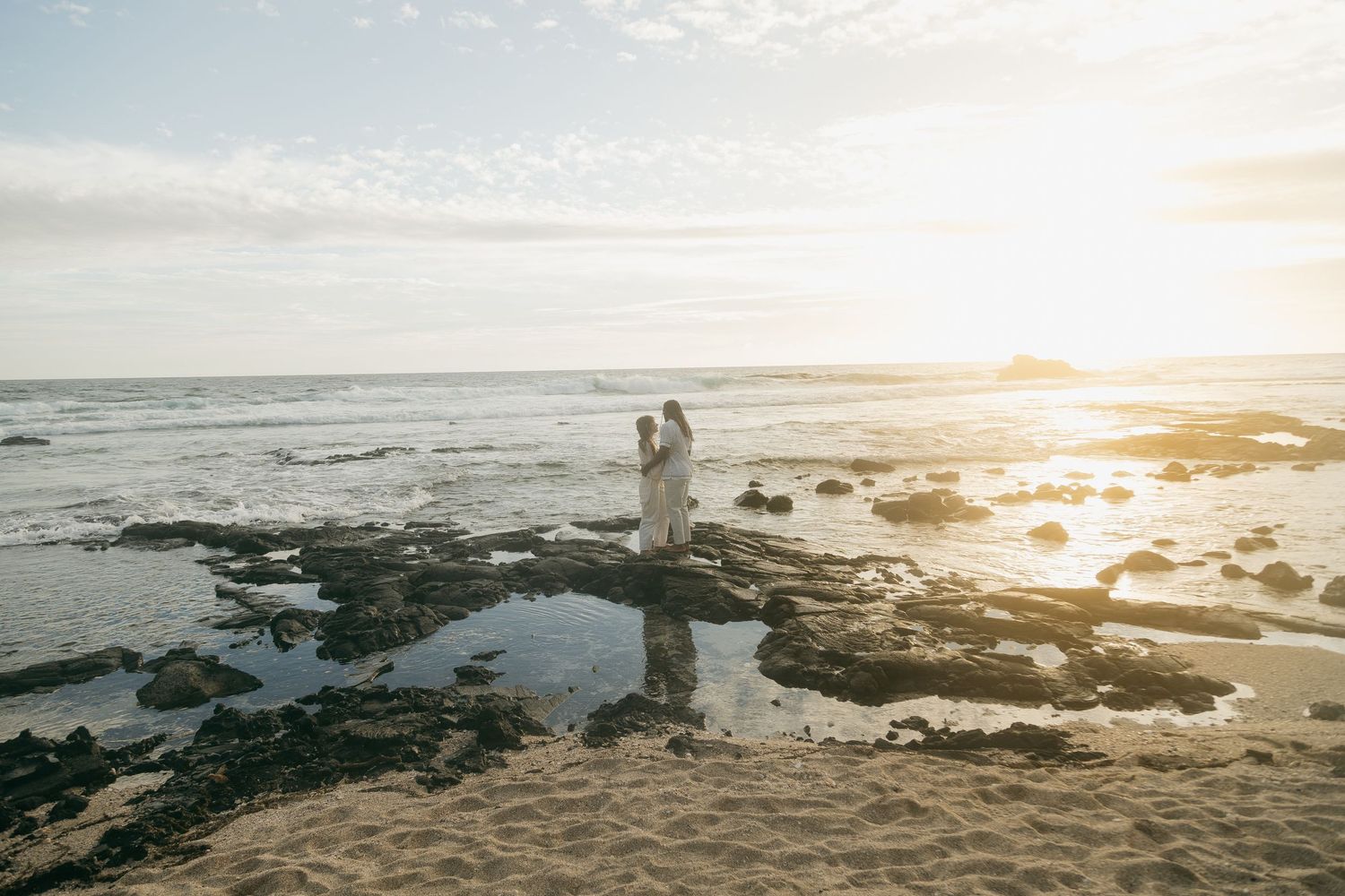 Couple walking along rocky beach coastline during golden sunset with waves crashing against shore.