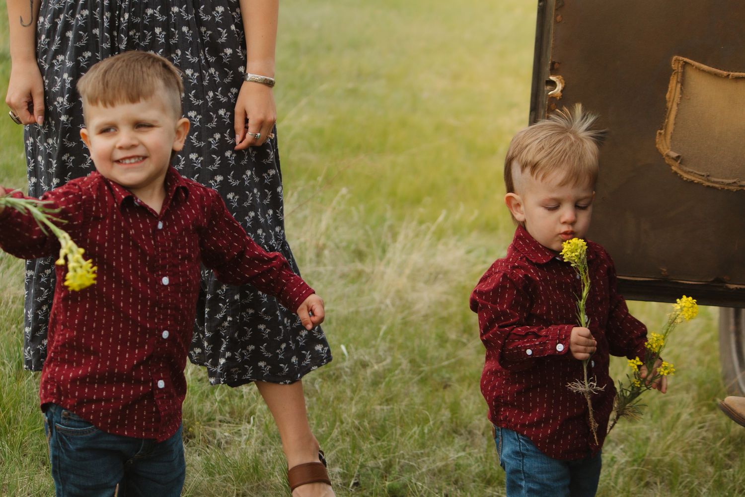Two children in matching red shirts holding yellow flowers while standing in a grassy field.