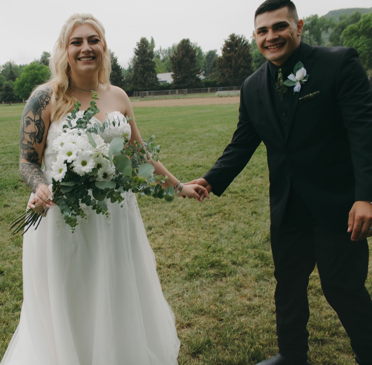 A cheerful wedding couple holding hands and bouquet while standing in a grassy field.