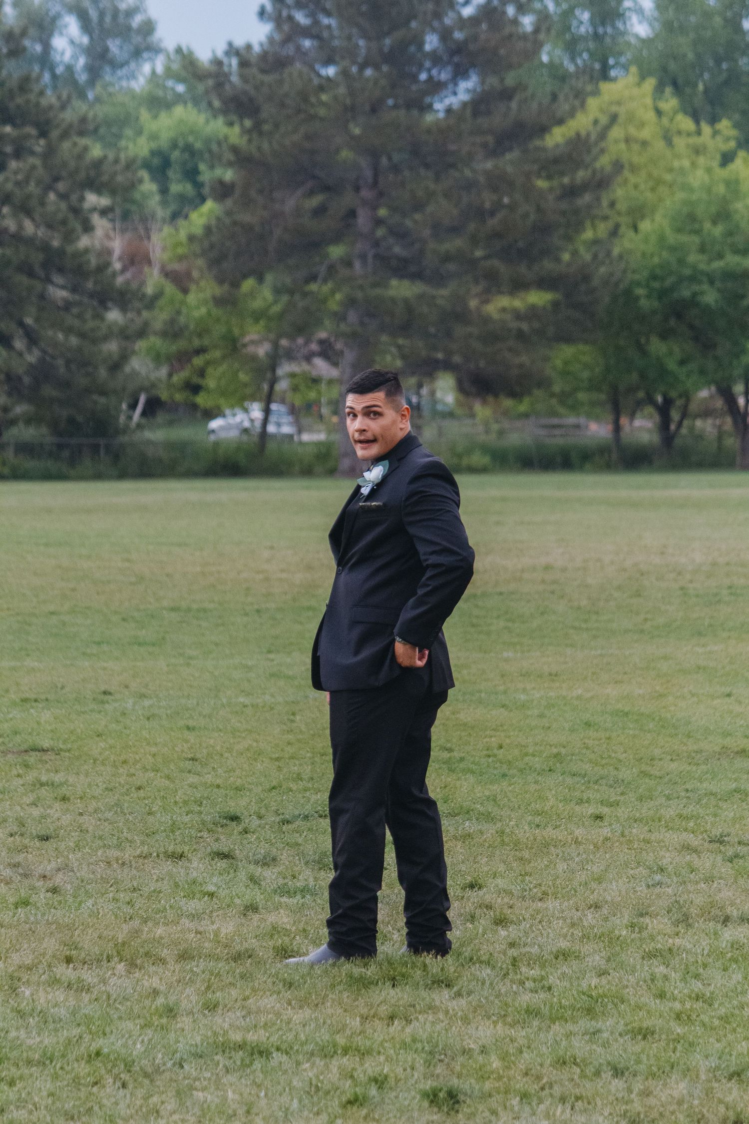 A groom in a classic black suit poses confidently in an outdoor setting with trees in background.