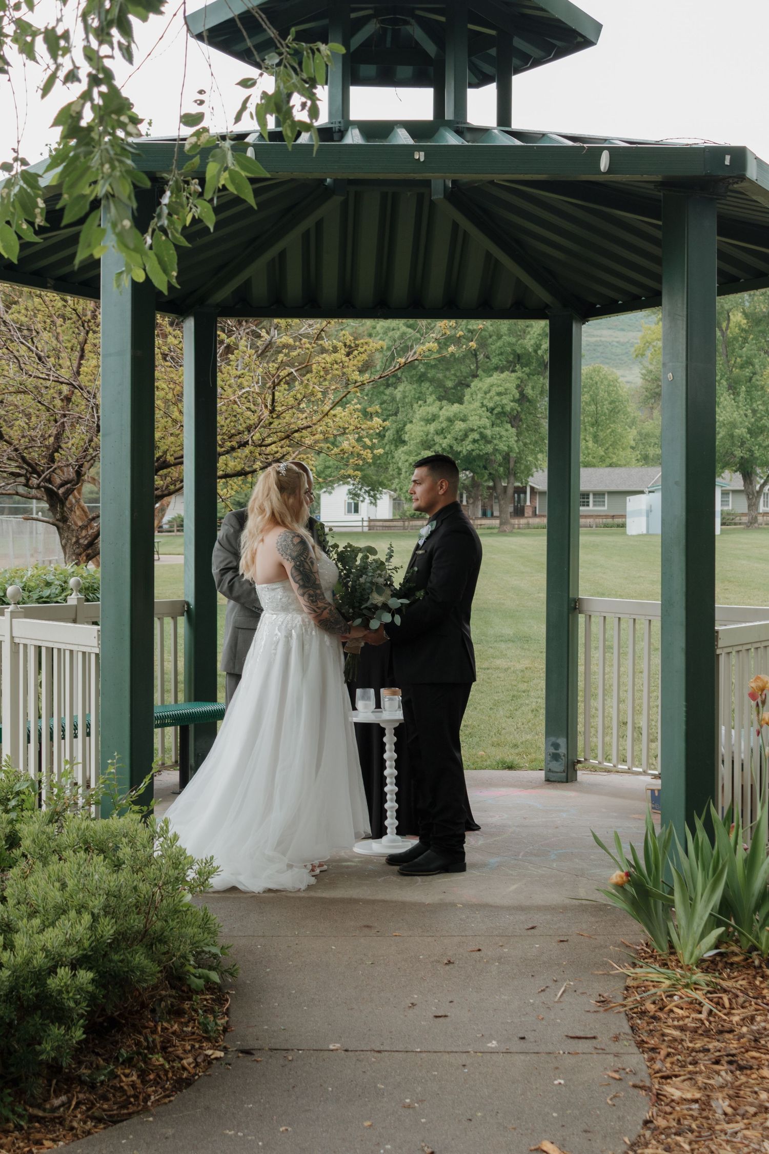 A couple exchanges vows in a green-roofed gazebo surrounded by spring foliage.