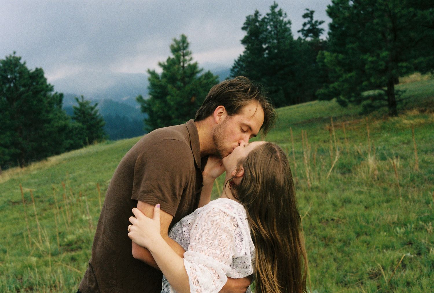 A couple sharing a romantic kiss in a lush green meadow with trees and cloudy mountains in the background.