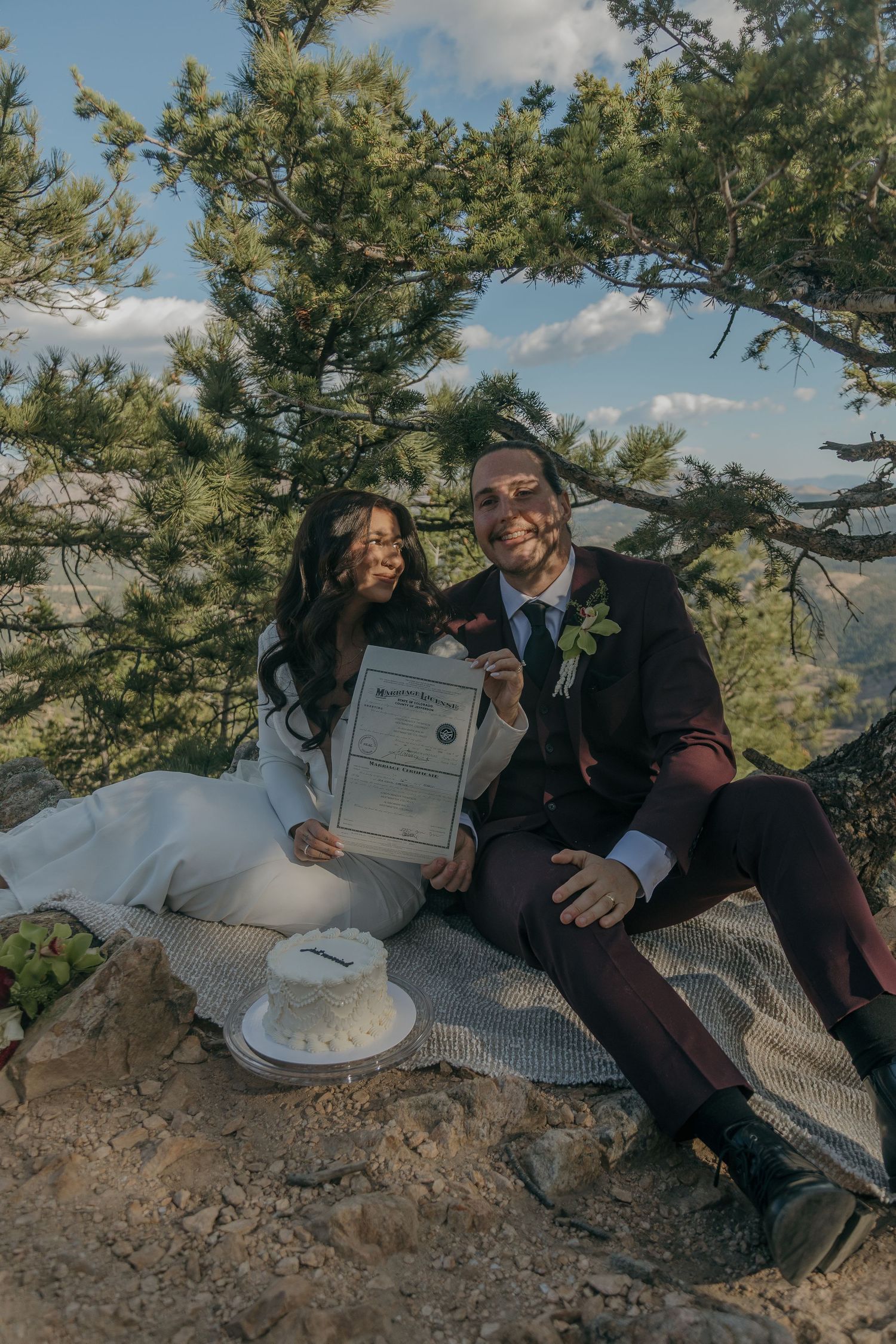 Newlywed couple sitting on rocky mountain overlook holding marriage certificate, bride in white gown, groom in burgundy suit.