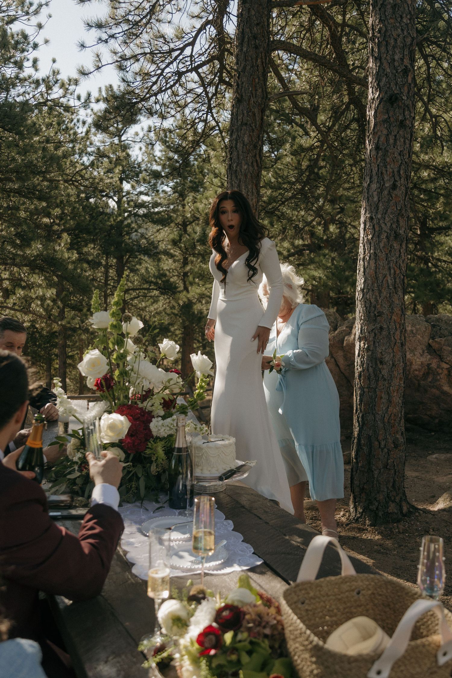 Bride in white gown standing at outdoor forest wedding table with floral decorations.