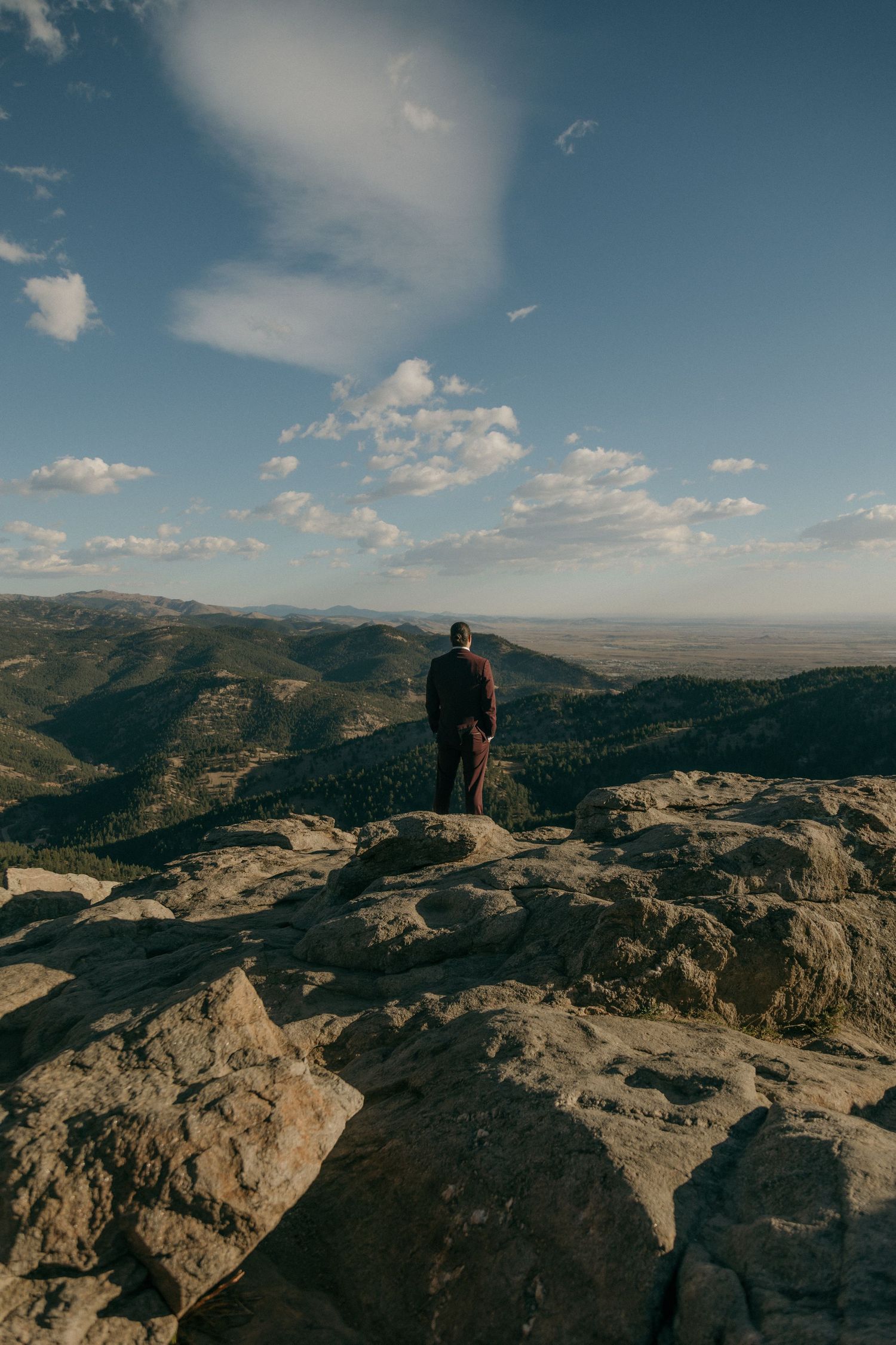 Groom in dark suit stands alone on rocky mountain summit overlooking vast Colorado valley under blue sky.