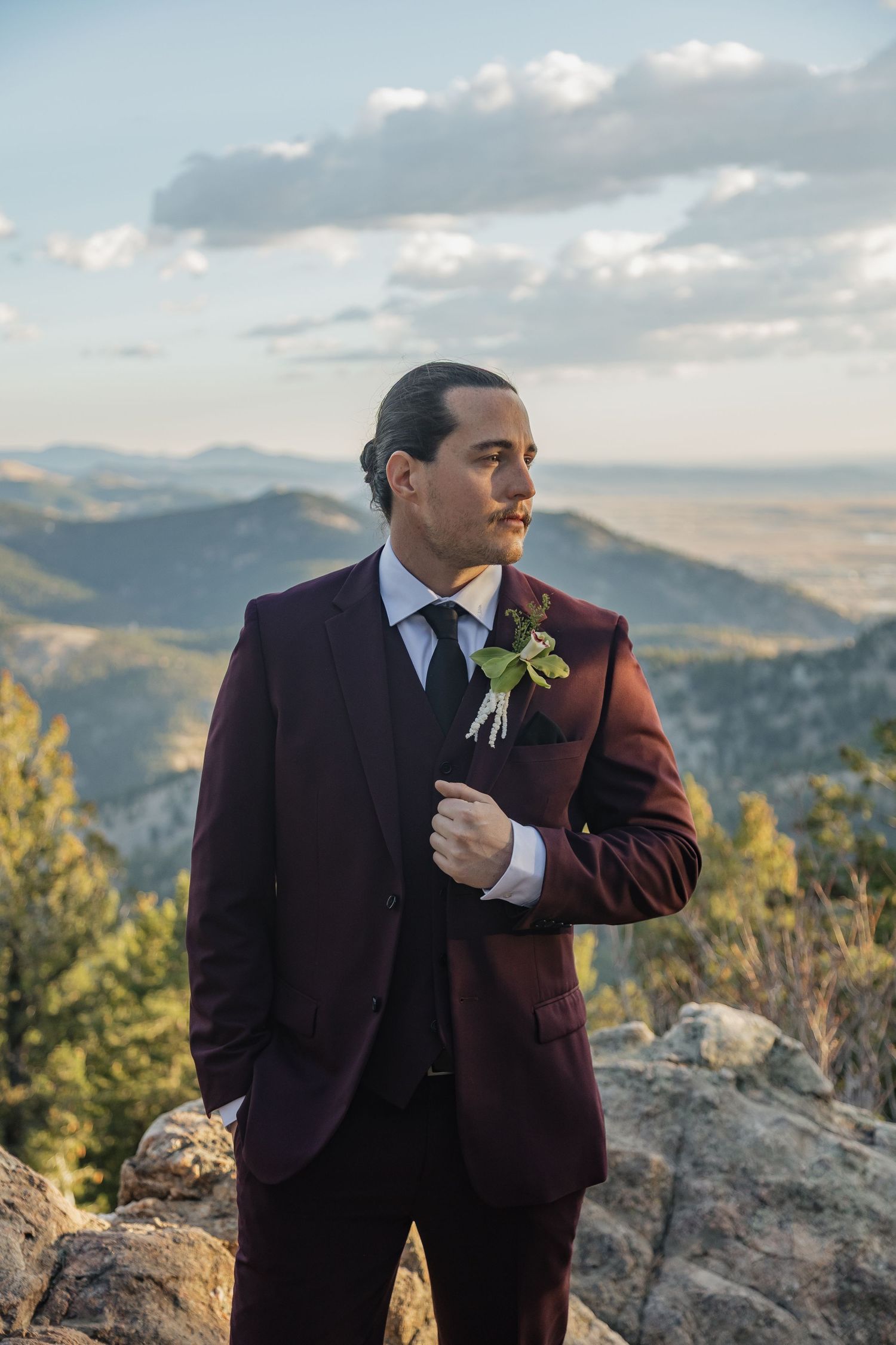 Groom in burgundy suit with boutonniere poses on rocky mountain overlook at sunset.