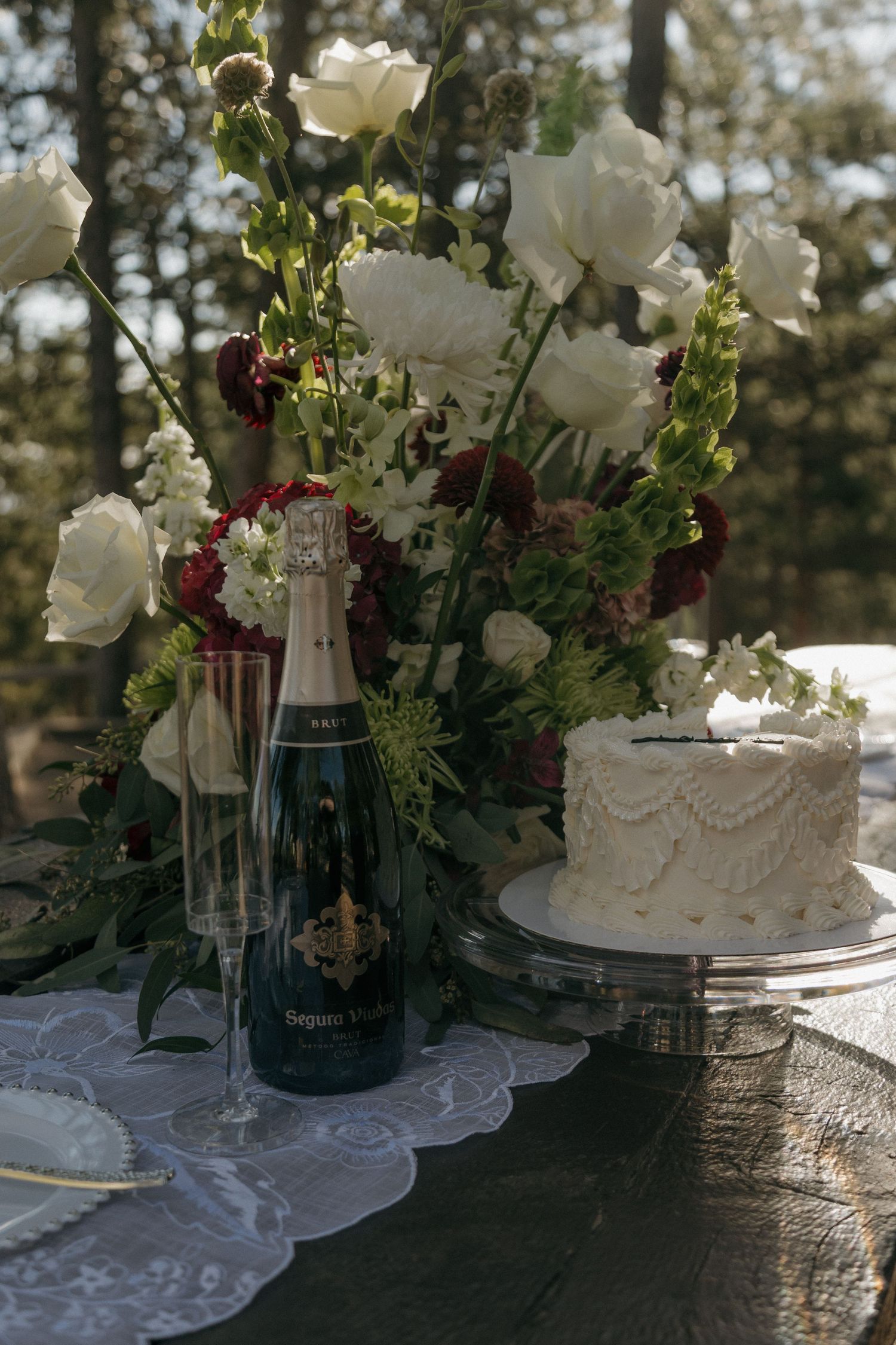 Elegant wedding table with white cake, champagne bottle, floral arrangement of white and burgundy blooms outdoors.