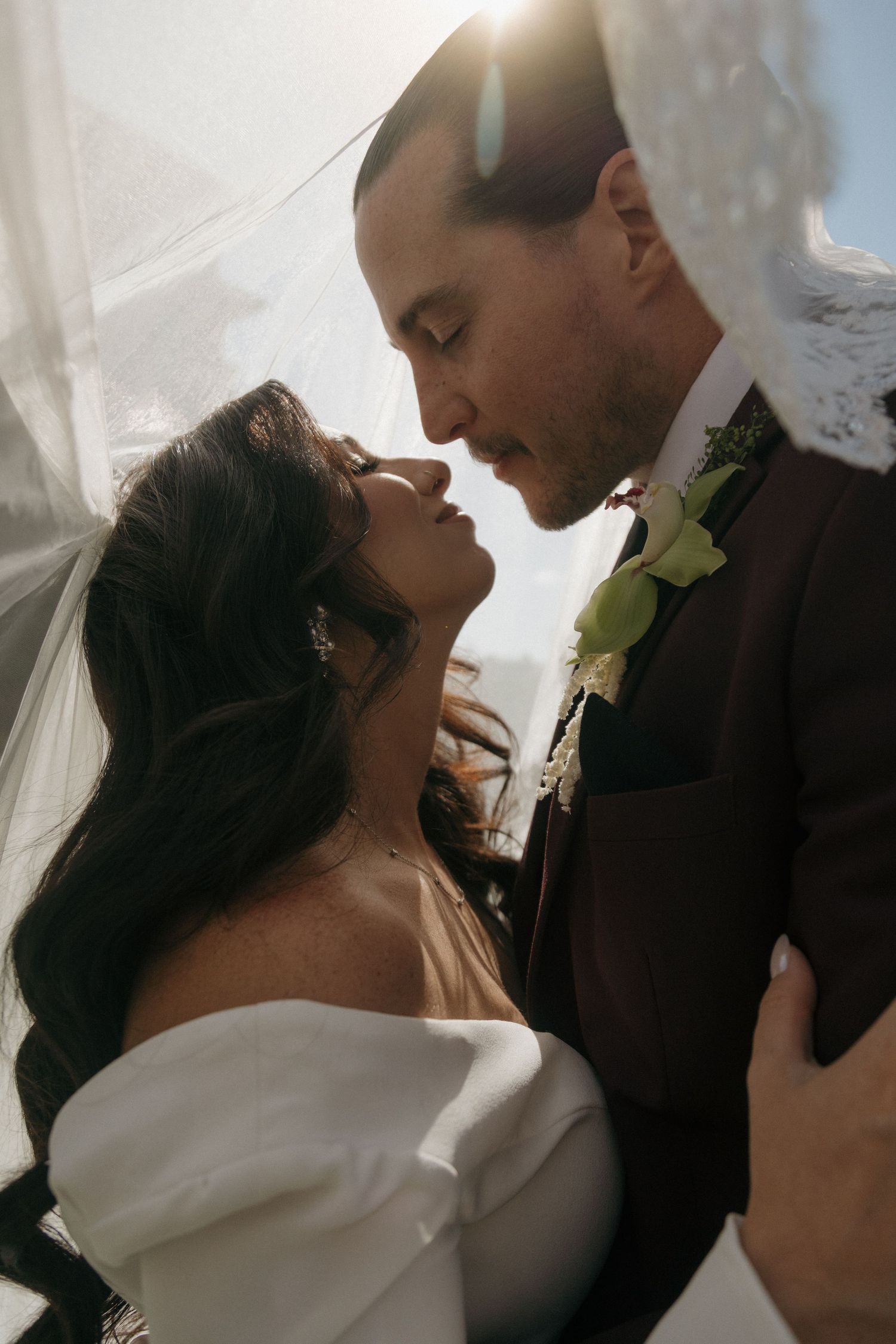 Bride and groom share an intimate moment under a flowing veil with soft backlit sunlight at their wedding.