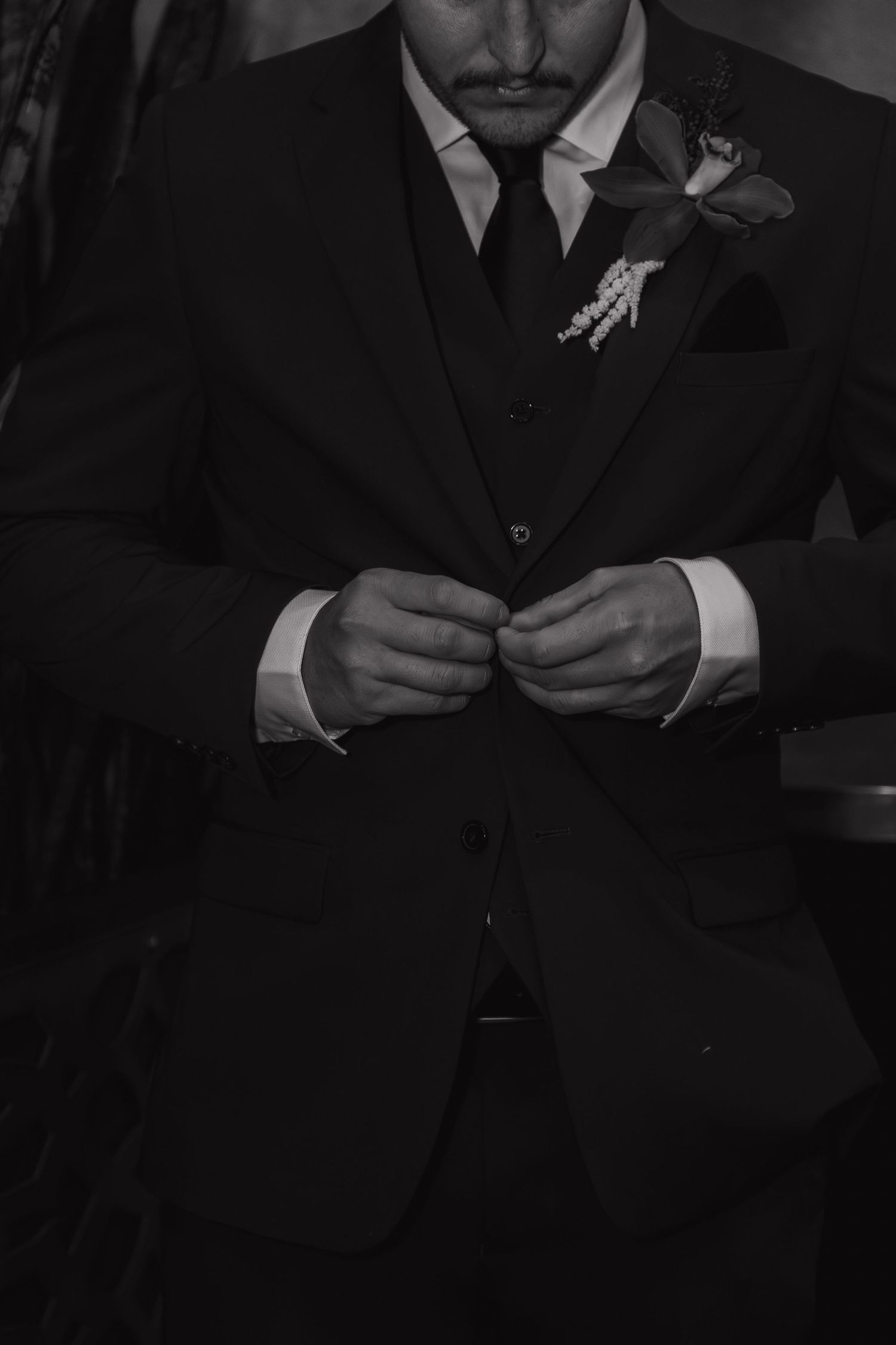 Black and white photo of a groom in a dark suit buttoning his jacket with a floral boutonniere.
