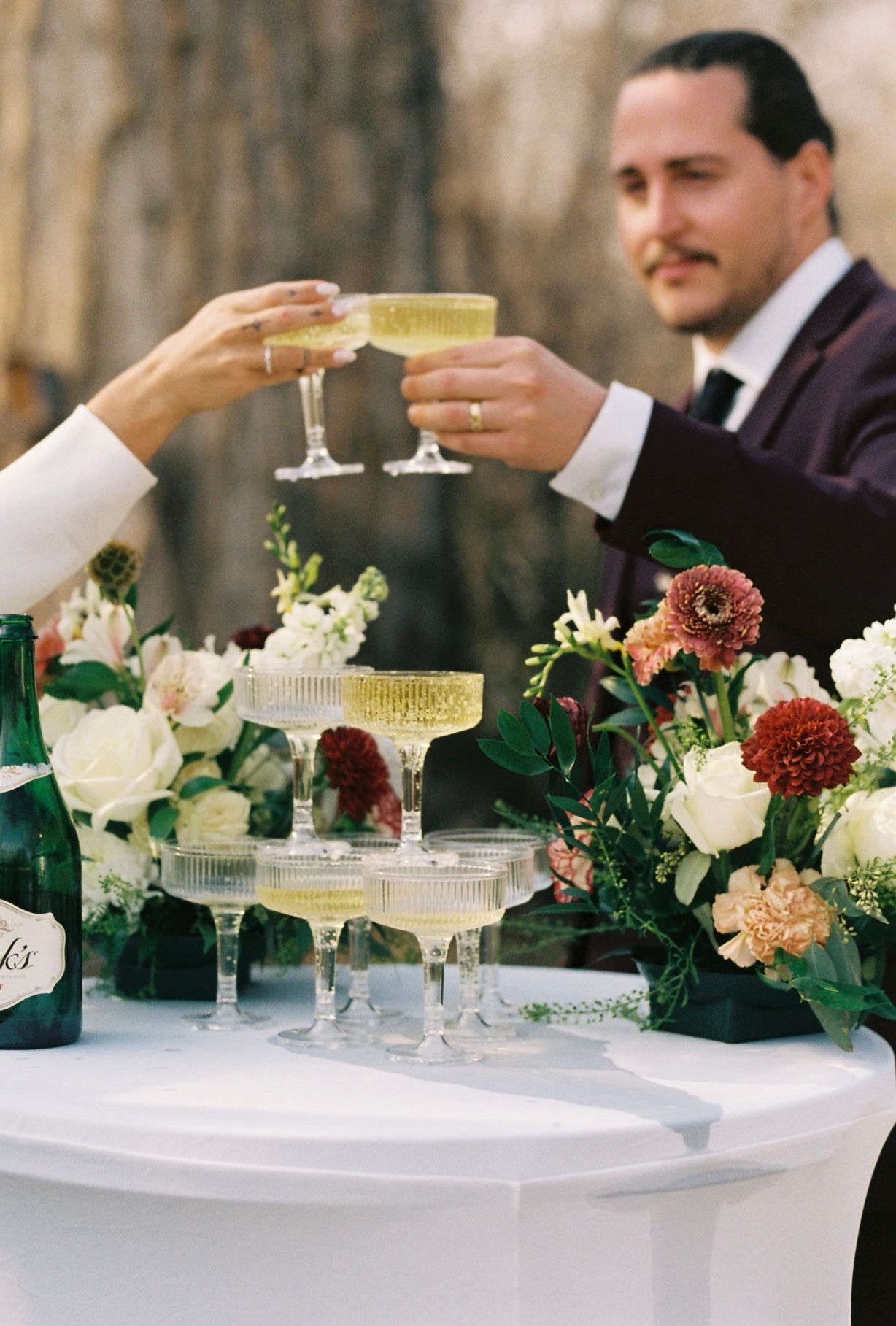 Bride and groom toasting champagne glasses over tiered coupe tower with floral arrangements.