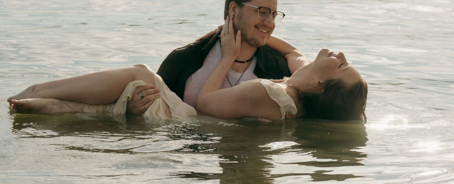 Two people in water, one supporting the other in a baptism or swimming lesson scene outdoors.