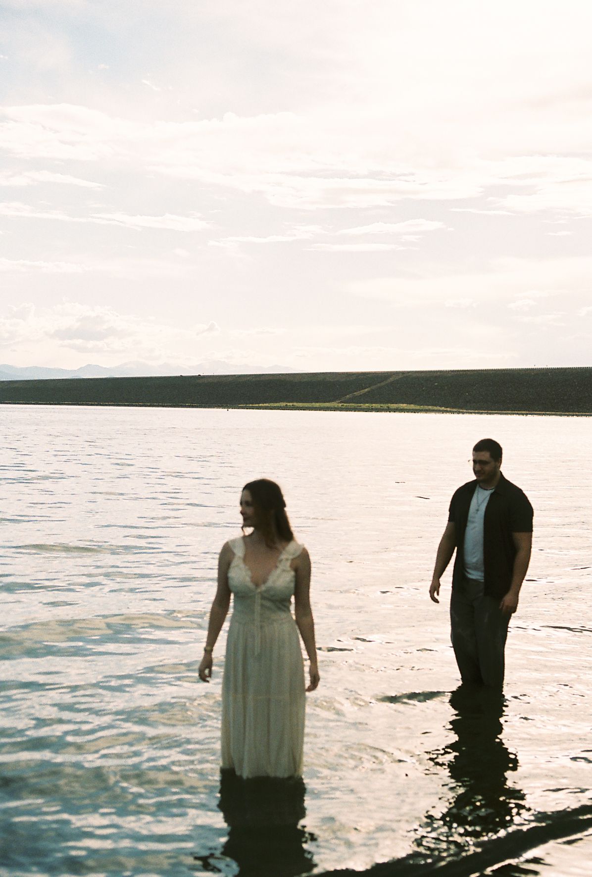 Couple wading through a calm lake at sunset with rolling hills in the background.
