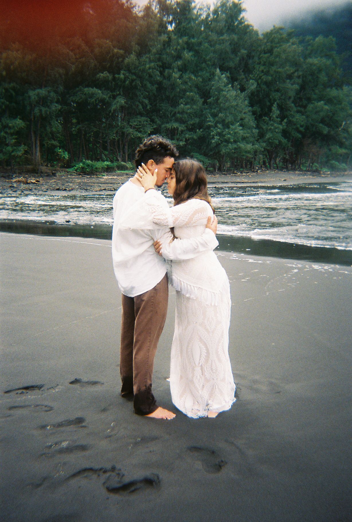 Couple embracing and kissing on a black sand beach surrounded by lush green trees and gentle waves.