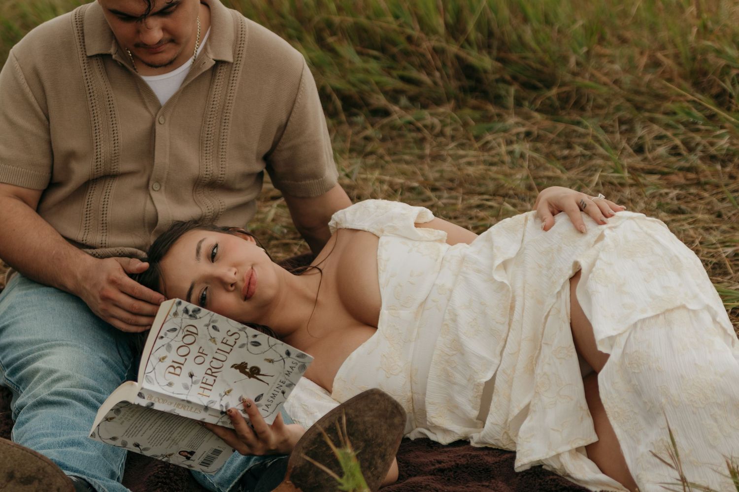 Couple relaxing outdoors, woman lying down reading a book while partner sits beside her in a field.
