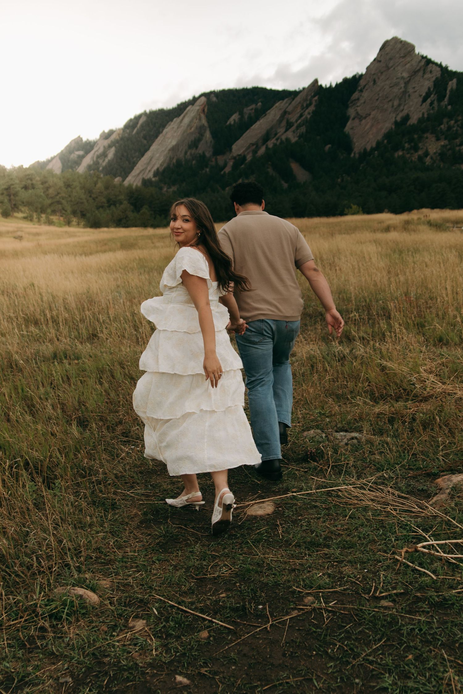 Bride in white tiered dress smiles back while walking with groom through golden meadow with Flatirons backdrop.