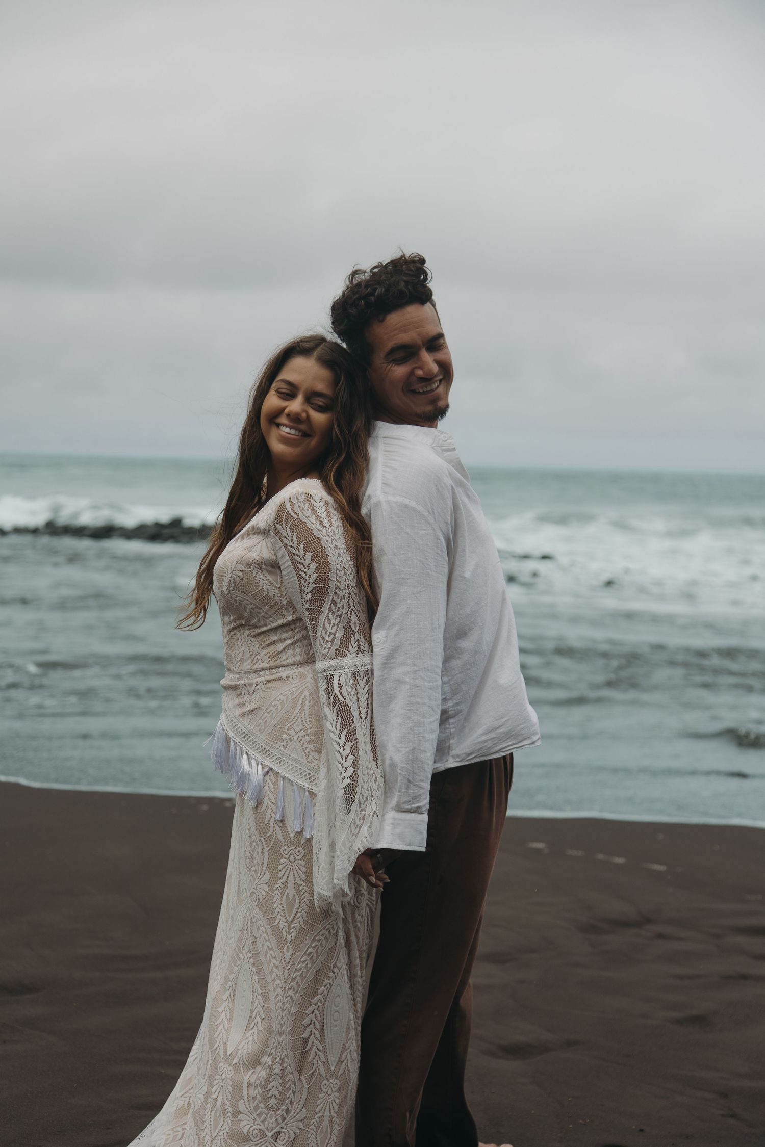 Smiling bride in bohemian lace dress and groom posing back-to-back on black sand beach by the ocean.
