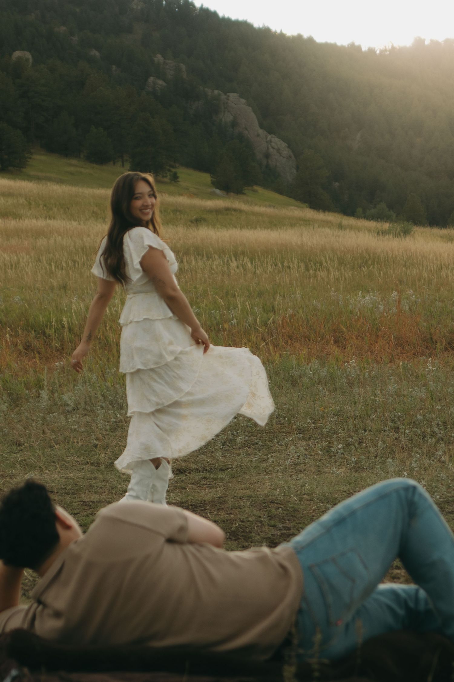 Smiling woman in white dress glancing back at partner lying in golden field at sunset.