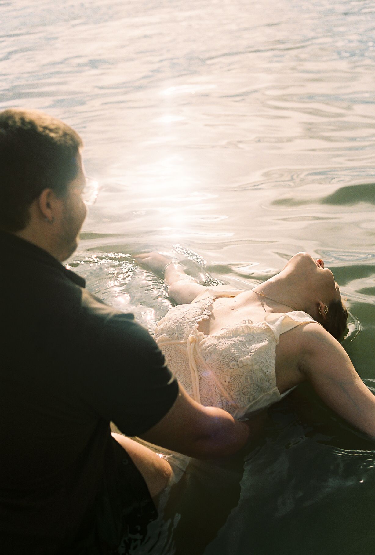 A bride in lace gown floats in sunlit water, cradled by a man in black suit during a romantic shoot.