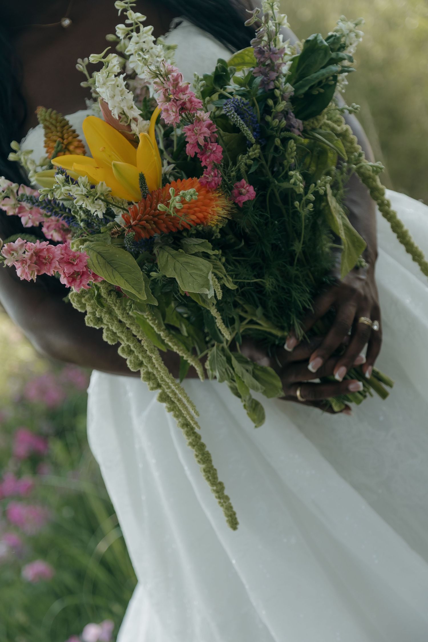 Black bride in white dress holding a lush wildflower bouquet with yellow, pink, and orange blooms in a garden.