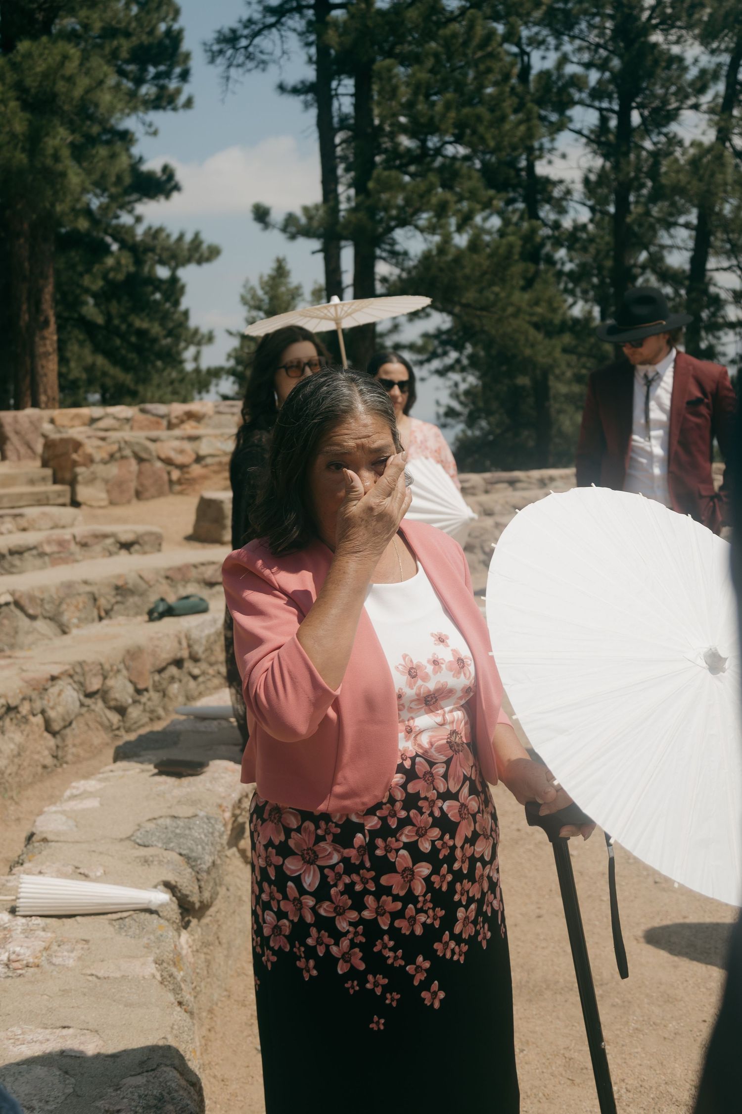 Emotional wedding guest in pink cardigan holds white parasol during outdoor ceremony among pine trees.