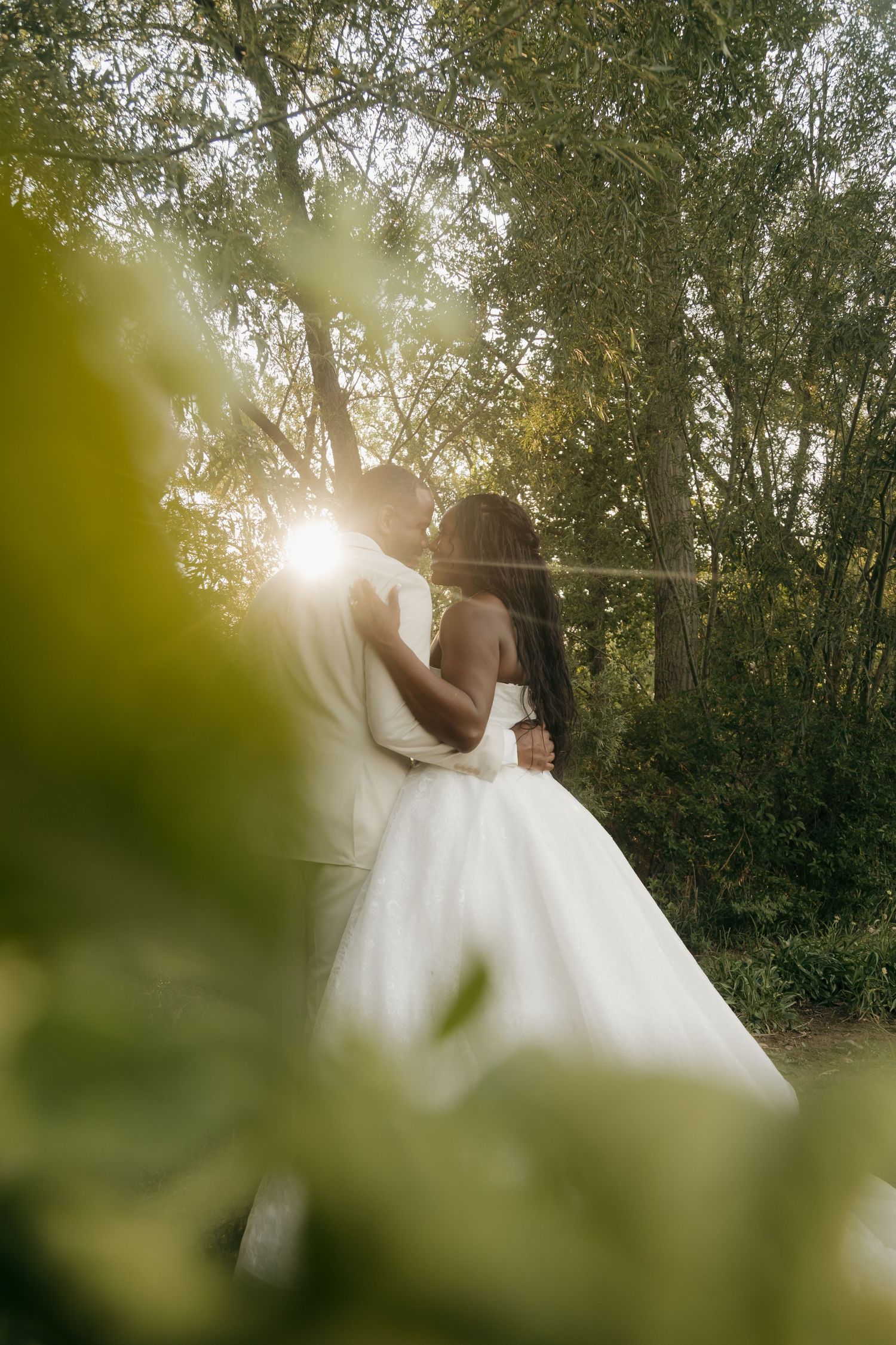 Bride and groom share a romantic kiss surrounded by lush green trees with golden sunlight glowing.