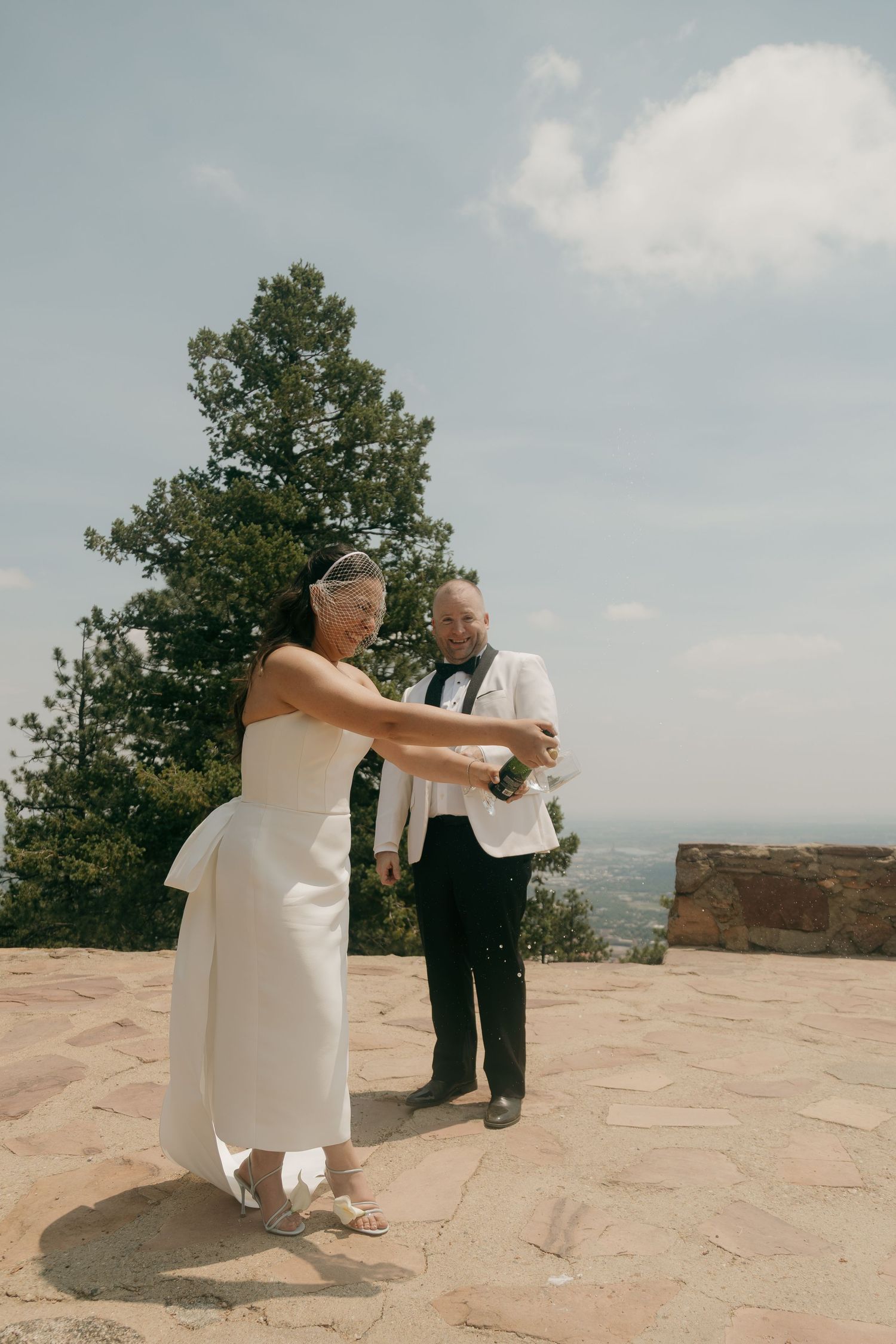 Bride and groom celebrate at mountain wedding venue, popping champagne bottle under blue sky.