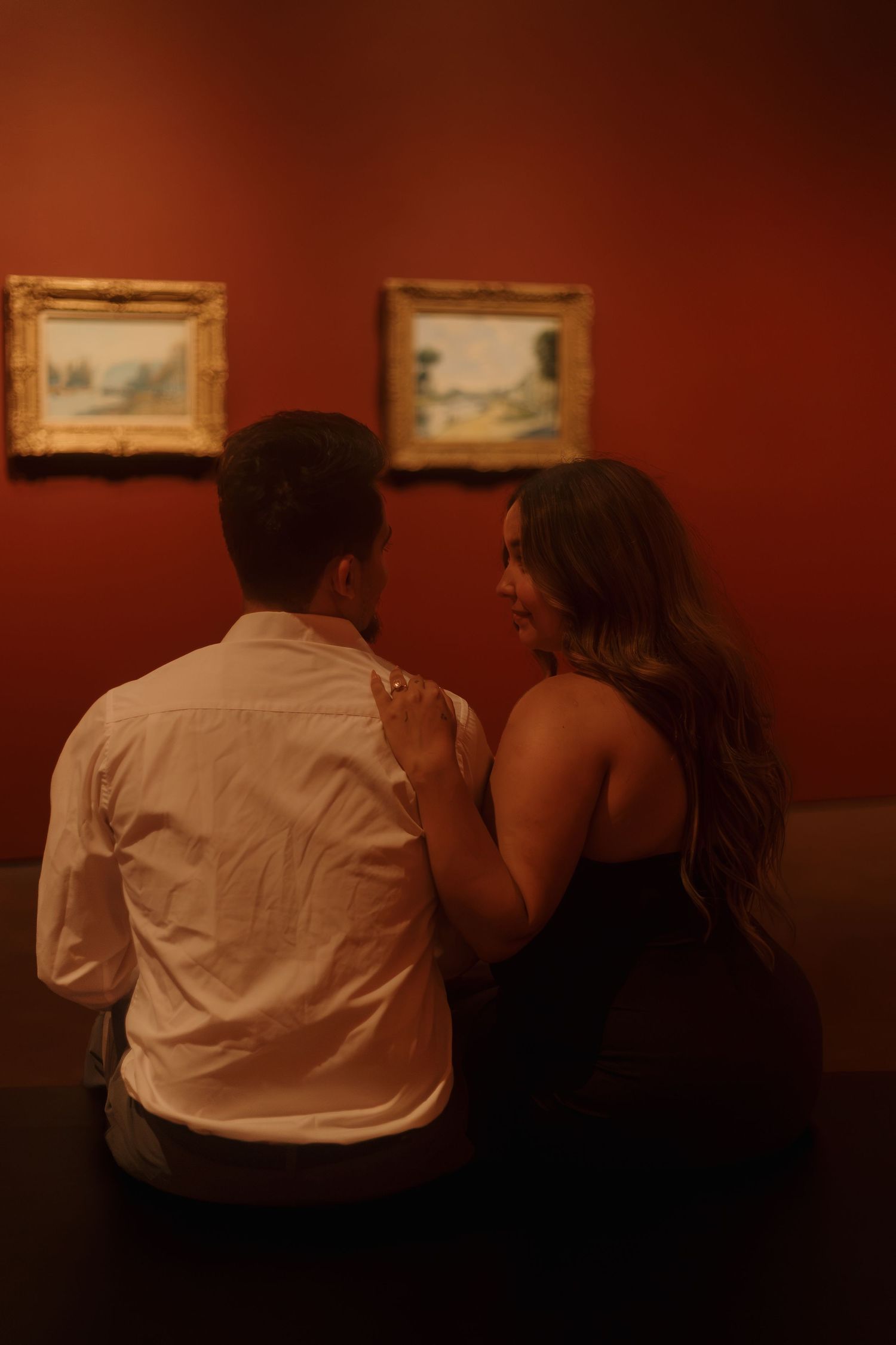Romantic couple sitting together on a bench in a warmly lit red museum gallery viewing landscape paintings.